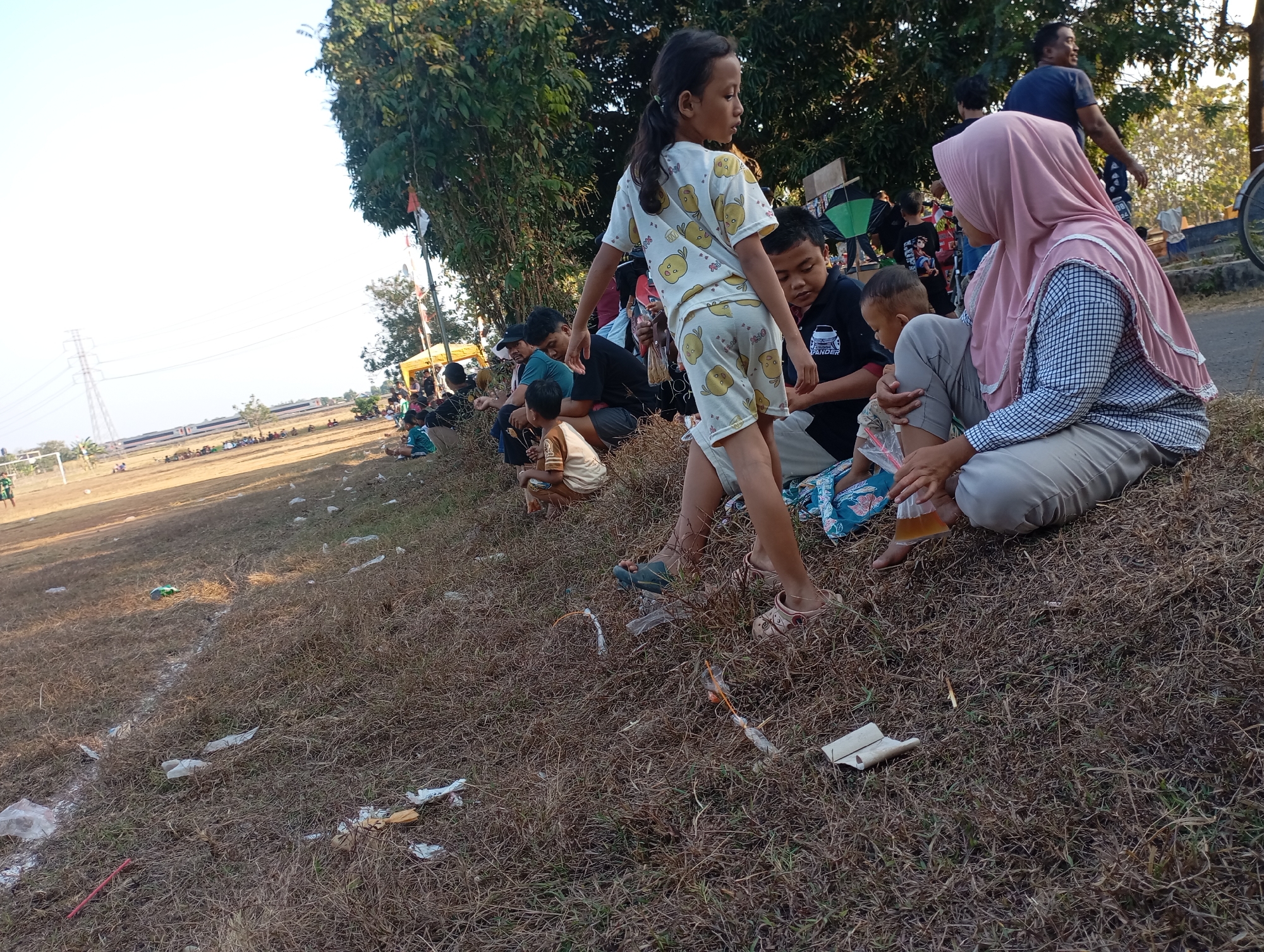 In Kecamatan Haurgeulis, Indonesia, on a dry, grassy field with scattered debris, a group of people, predominantly children and adults, are gathered. A young girl in a yellow shirt with cartoon prints stands in the foreground, looking to her right. Behind her, several individuals are seated, including a woman wearing a pink hijab and a checkered shirt. A young child is being held by an adult to her right. In the background, more people are visible, some sitting and others standing, with trees and a utility pole in the distance. The ground is covered in dry, brown grass with some white lines visible, possibly marking a field. There are various pieces of trash scattered on the ground, including plastic bags and wrappers. The overall scene suggests an outdoor gathering or event.