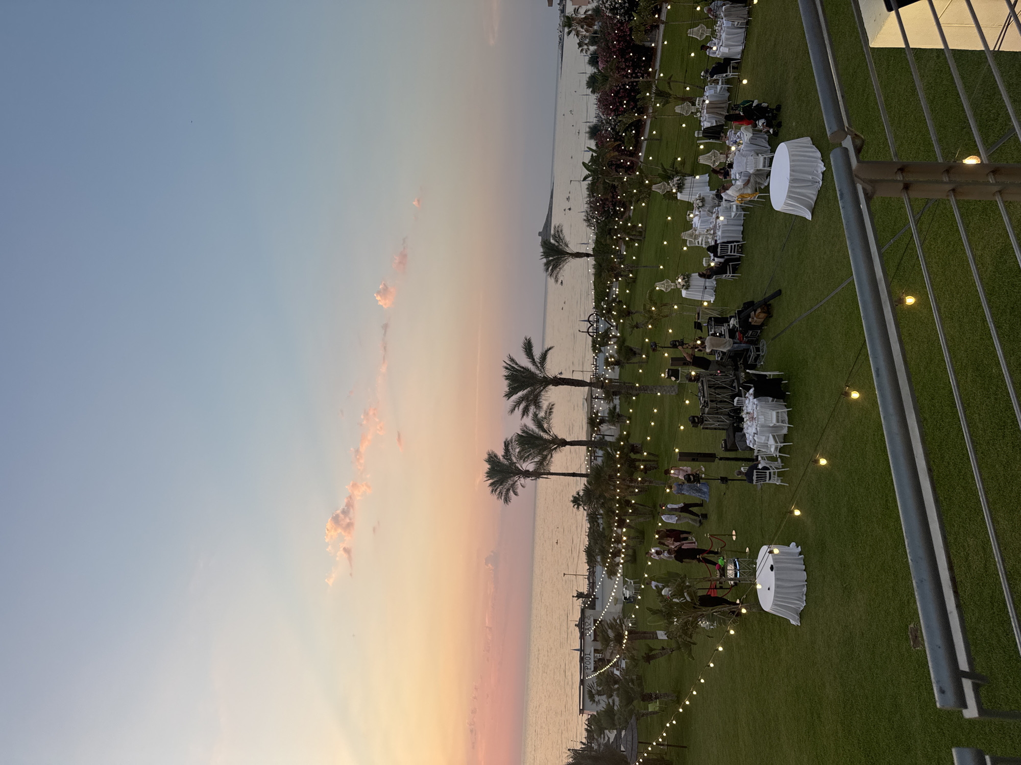 This image depicts an outdoor event, likely a wedding reception or formal gathering, set against a scenic coastal backdrop during sunset. The scene is viewed from an elevated perspective, looking down onto a meticulously maintained green lawn where numerous round tables, draped in white tablecloths and adorned with white chairs, are arranged. Strings of warm, glowing Edison-style bulbs are strung across the lawn, illuminating the space and creating a festive ambiance.
Several people are visible, mostly seated at the tables or standing and mingling. Their attire suggests a formal occasion, with many guests dressed in dark clothing. In the background, palm trees and other lush vegetation line the edge of the lawn, separating it from the calm, shimmering water of the sea. The sky transitions from a soft blue at the top of the frame to warm hues of pink, orange, and yellow near the horizon, indicating it is either sunrise or sunset. A few wispy clouds catch the light, adding texture to the sky.
The presence of what appears to be a small stage or setup with lighting equipment and a camera suggests entertainment or a performance is part of the event. The overall mood is serene and elegant, enhanced by the natural beauty of the location and the warm, inviting lighting. No visible text is present in the image. The location context of Isola delle Femmine, Italy, reinforces the coastal setting and suggests a Mediterranean atmosphere.
