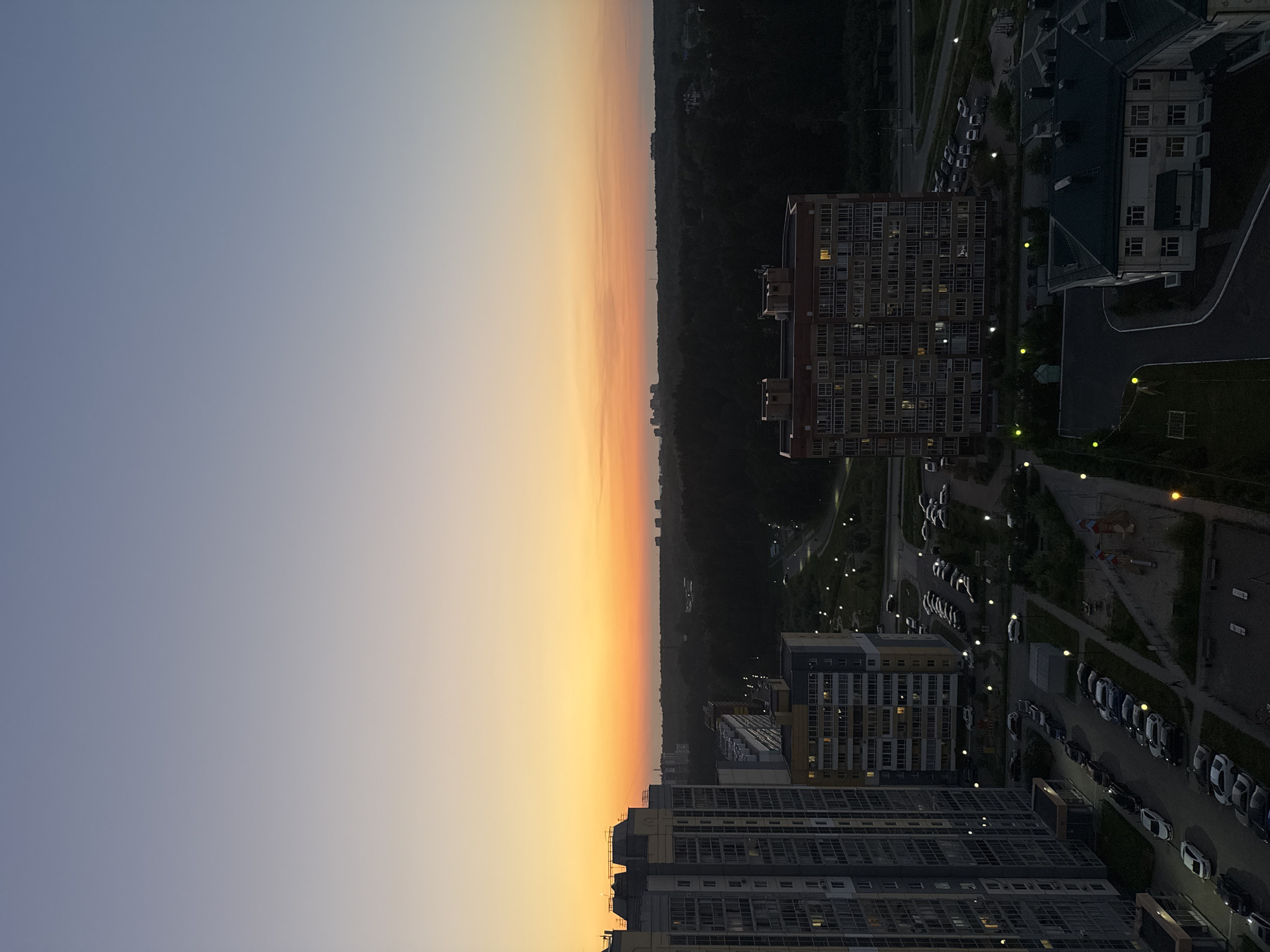 The image depicts a high-angle view of a residential area at dusk, likely in Tomsk, Russia. The sky transitions from a clear, pale blue at the top to a warm orange and yellow gradient near the horizon, suggesting sunset. Silhouetted against the sky are several multi-story apartment buildings. In the foreground, a tall, grey and white residential building dominates the left side of the frame. To its right, another apartment building with yellow and grey accents is visible. Further into the background, a dense, dark forest stretches across the landscape, with distant, smaller buildings appearing as dark shapes. The ground level shows parking lots filled with cars, streetlights illuminating pathways, and glimpses of playgrounds and other amenities. The overall atmosphere is calm and quiet, characteristic of a residential neighborhood settling down for the evening.