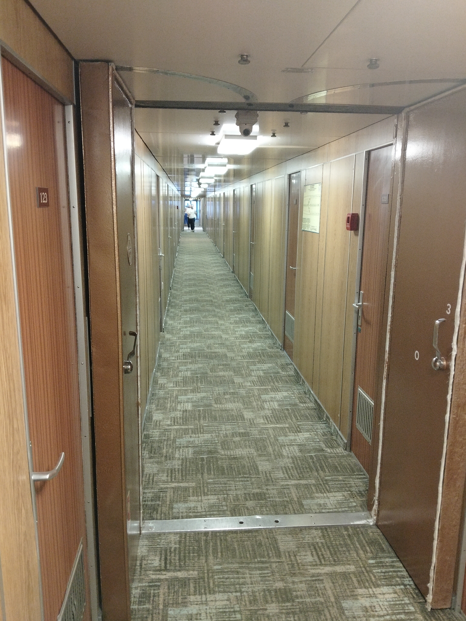 The image shows a long, narrow hallway on a ship or train, lined with identical doors. The hallway is carpeted with a patterned gray and green carpet. The doors are made of wood and have metal handles. There is a sign on one of the doors with the number 123. At the far end of the hallway, a person can be seen walking away from the camera. The lighting in the hallway is provided by fluorescent lights in the ceiling. The overall impression is one of a functional and utilitarian space. There is no indication of the location being in the city of Kozmodemyansk, Russia.