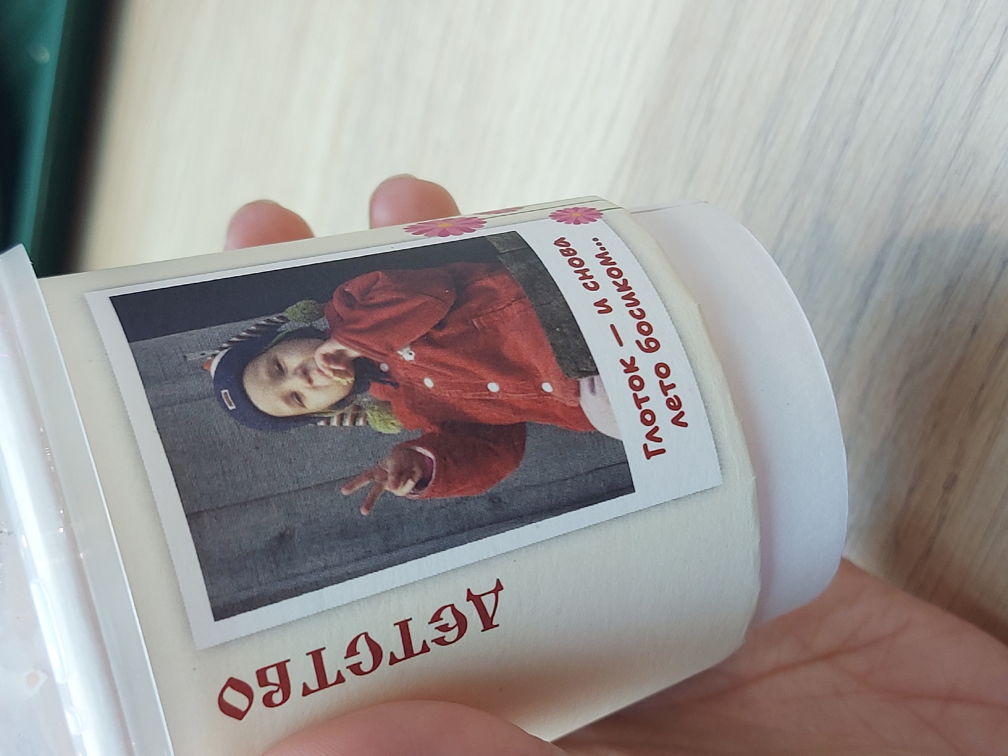 A close-up shot shows a white container, presumably a jar or cup, held in a person's hand. A printed label is adhered to the container, featuring a photograph of a child wearing a red jacket and a blue hat, making a peace sign with their hand. The photo has a white border reminiscent of a Polaroid print. Cyrillic text appears on the label: "ДЕТСТВО" at the bottom and "ГЛОТОК — И СНОВА ЛЕТО БОСИКОМ..." above the picture. The background of the image is a light-colored surface with a wood grain pattern. Another person's fingers are visible in the background, as is a hint of something green. The text suggests a Russian origin and context. Without more information, a specific location in Saint Petersburg cannot be determined from the image.