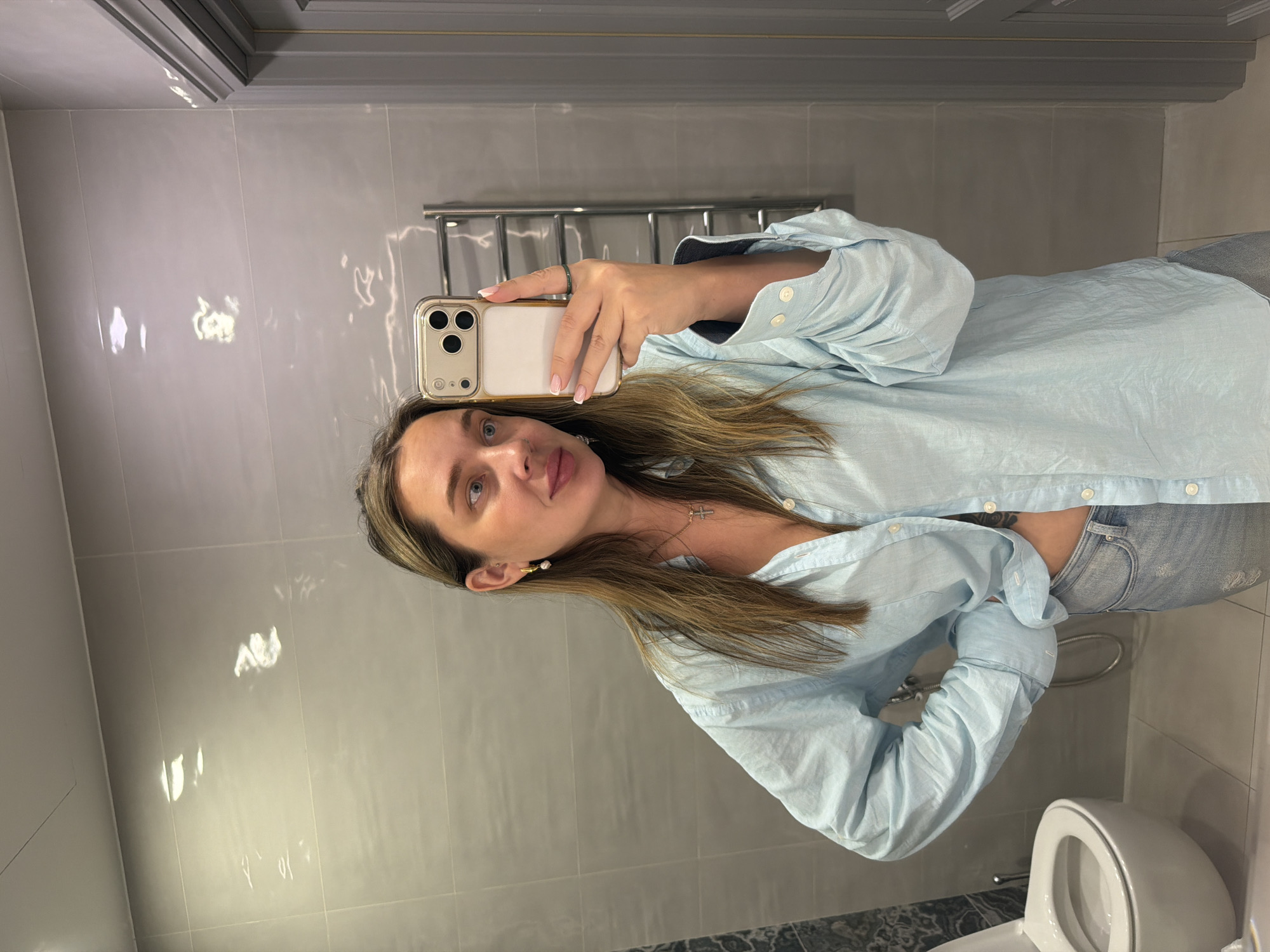 A woman is taking a selfie in a bathroom with gray tile walls. She is wearing a light blue button-down shirt and jeans. She is holding a gold-colored iPhone. A toilet and towel rack are visible in the background. There is no indication of the city of Moscow, Russia.
