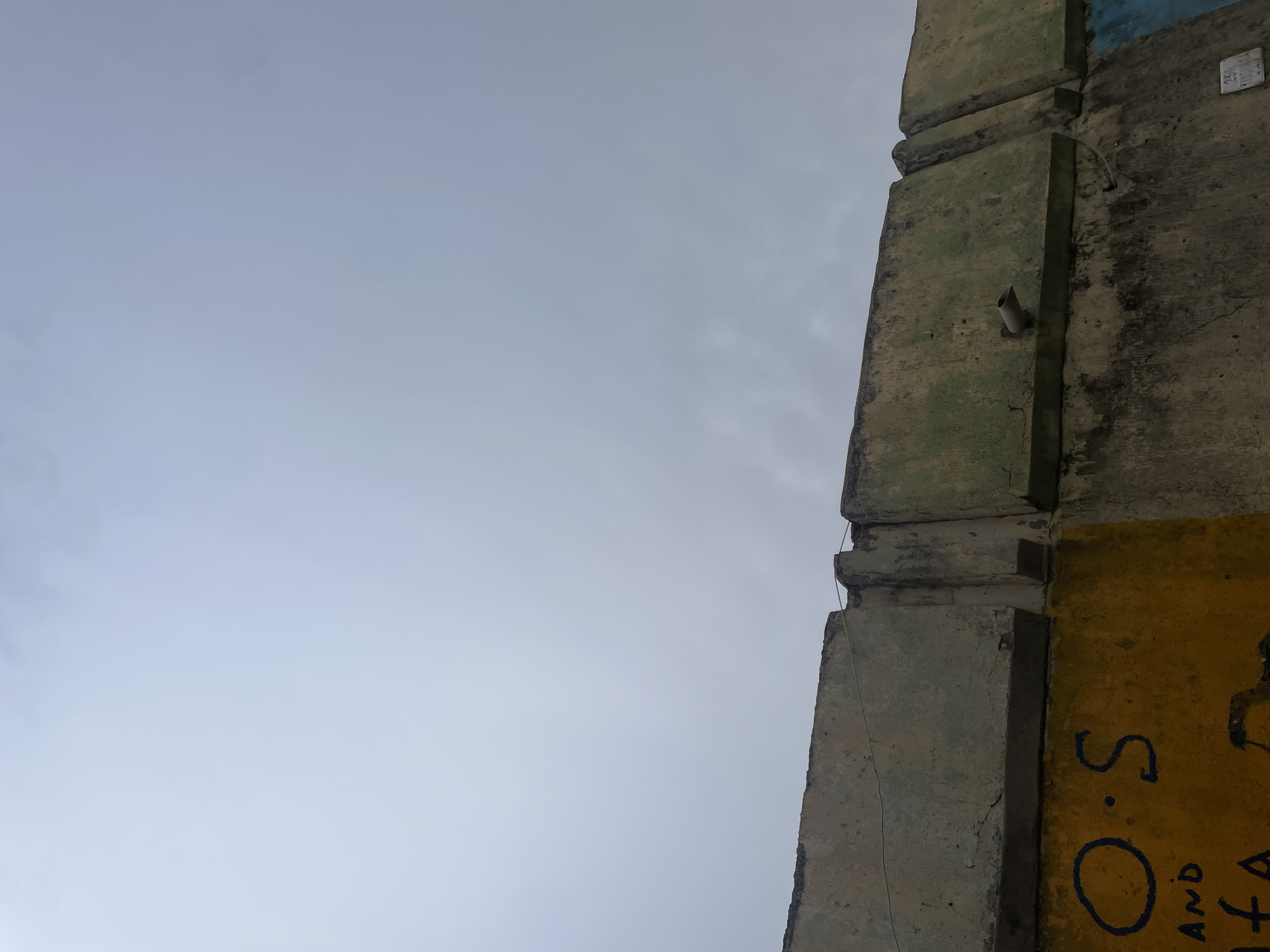 The image displays a diagonal composition, with a weathered concrete structure on the right and an expanse of overcast sky on the left. The concrete structure, possibly a pillar or wall, is made of stacked, rectangular blocks and shows signs of age with grey and greenish discoloration, cracks, and rough textures.
Near the top right, a small white rectangular plaque is affixed to the concrete. It contains the numbers "258" and "008", followed by text that appears to be "कि० मी०" (likely "kilometer" in Hindi). Below this plaque, a short, white, angled pipe or conduit protrudes from the concrete. A thin wire or string runs vertically along the structure's edge.
Towards the bottom right, a section of the concrete wall is painted yellow. On this yellow surface, large black letters are visible as graffiti, spelling out "S. O." followed by "AND" directly beneath it, and then "TA", which appears to be part of a larger, incomplete word.
The sky occupies the majority of the left side of the frame, appearing a uniform light grey, indicating cloudy or overcast weather during the daytime. There are no people or specific activities taking place. The scene is static, capturing an architectural element against the sky, possibly in an urban environment.