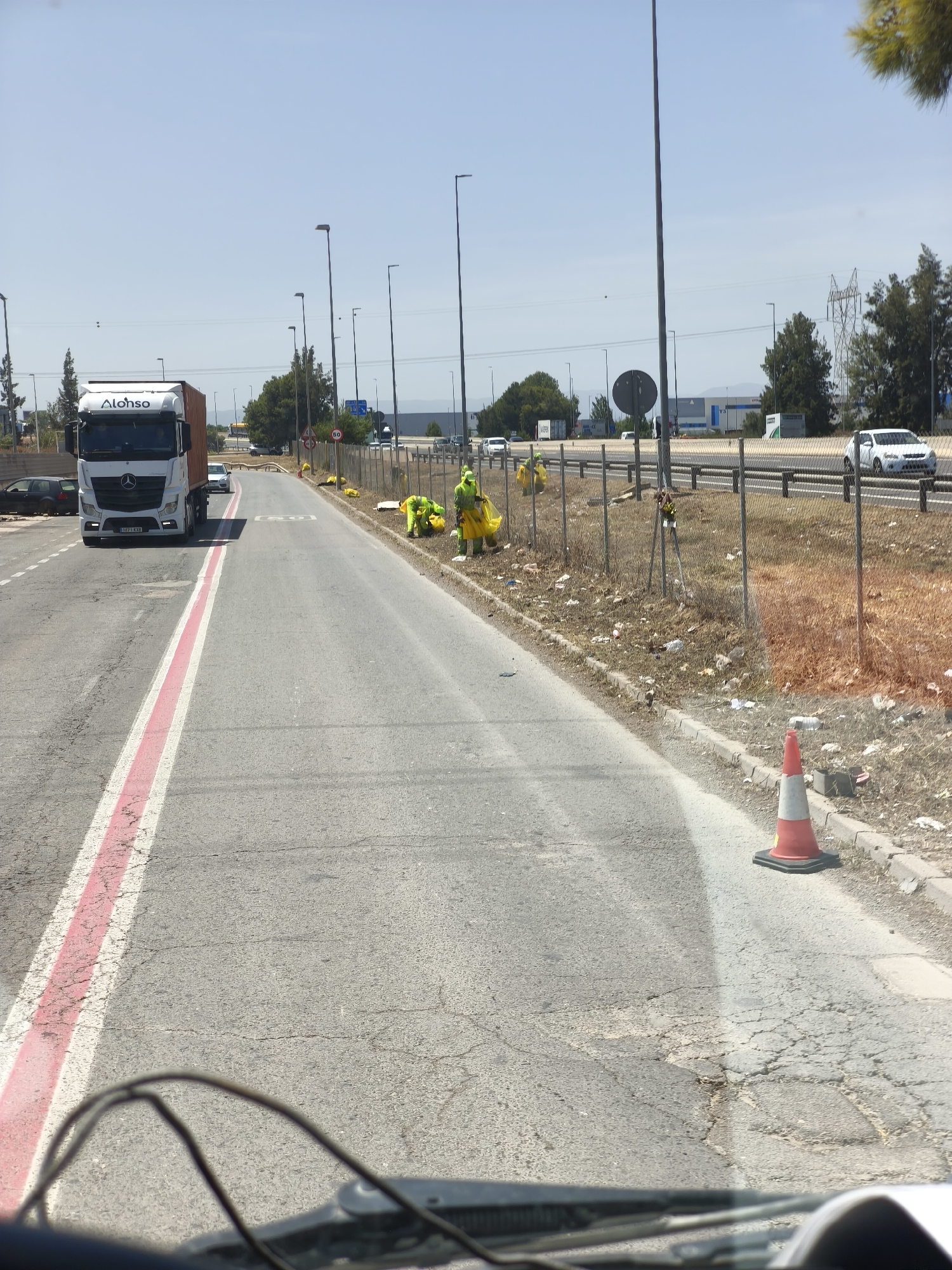 The image captures a sunny daytime scene along a roadside in Quart de Poblet, Spain, viewed from inside a vehicle. A clear blue sky dominates the upper portion. On the left, an asphalt road features a distinctive red and white painted line. A large white Mercedes-Benz semi-truck, bearing the name "Alonso" on its cab and license plate "3187 KXB", is driving away from the viewer in this lane.
To the right of the road, a dry, unkempt verge runs alongside a metal chain-link fence. Beyond this fence is a multi-lane highway with several cars visible, including a white car in the mid-ground. An "80" speed limit sign is noticeable near the highway.
At least three individuals, wearing bright yellow high-visibility suits, are actively engaged in roadside cleanup along the verge next to the fence. They appear to be collecting litter, with one holding a yellow bag. The verge itself is strewn with debris and dry vegetation. In the foreground on the right, a red and white traffic cone stands on the curb. Tall streetlights line both the immediate road and the distant highway. Industrial buildings and scattered trees are visible in the background, along with power lines and utility poles. The asphalt in the foreground shows signs of cracking and wear.