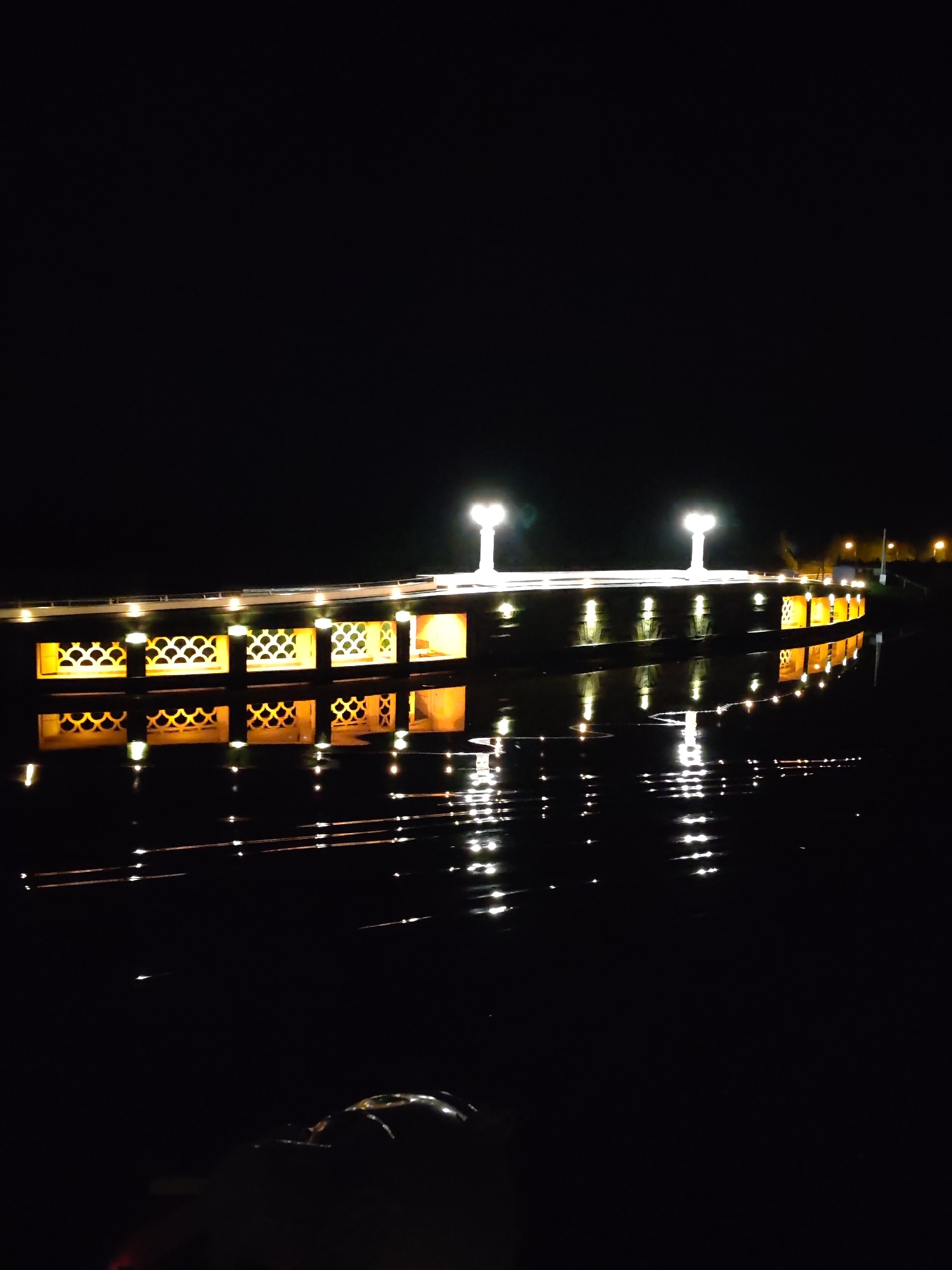 The image depicts a pier or structure extending over water at night. The structure is illuminated with evenly spaced lights along its length and two prominent lamp posts near the center. The lights cast reflections on the water's surface. The structure has decorative openings. The scene is set in a dark environment. There is some activity on the end of the structure. The location is Karmanovo, Russia.