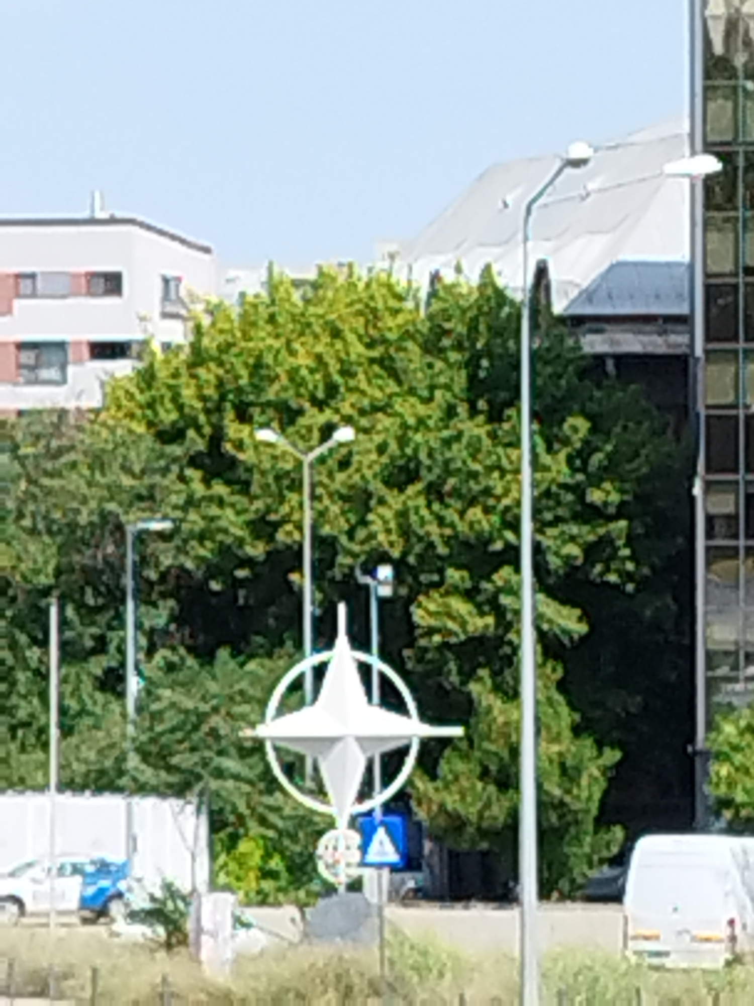 The image depicts an outdoor scene with a clear sky and some buildings in the background. In the foreground, a prominent white NATO star logo on a pole is visible, along with streetlights. Behind the logo, lush green trees form a dense canopy. To the left of the trees, a white building with several windows can be seen. To the right, a modern glass-fronted building dominates the background. A white van is parked on the right side of the image. On the left, a white car with blue accents is parked. The scene appears to be a public space, likely in an urban environment. While the precise location is not confirmed within the image itself, this particular NATO star installation is located at NATO's Supreme Headquarters Allied Powers Europe (SHAPE) in Mons, Belgium, not Bucharest, Romania.