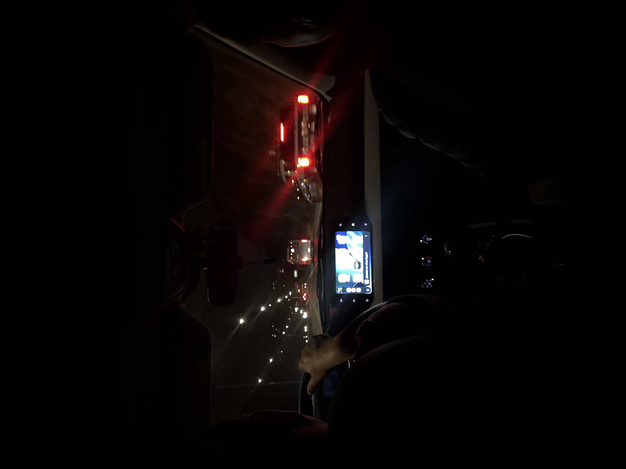 The image shows the interior of a vehicle at night, seemingly in slow-moving traffic in San Francisco Totimehuacán, Mexico. The vehicle is stationary or moving slowly, as evidenced by the illuminated brake lights of the car directly ahead, visible through the windshield. The driver is holding the steering wheel. A smartphone mounted on the dashboard is turned on, its screen displaying application icons. The dashboard controls are partially visible to the right of the phone. The overall ambient light is low, with the primary illumination coming from the external traffic lights and the smartphone screen.