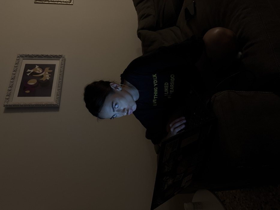 The image captures an indoor scene in a dimly lit room, located in the city of Venice, Italy. A female individual, appearing as a young adult, is seated on a dark-colored upholstered sofa, positioned slightly off-center to the right, looking directly at the camera. She wears a dark long-sleeved shirt with the partially visible yellow text "EVERYTHING YOU LIKED FIVE YEARS AGO." Her dark hair is pulled back, and her face is illuminated by the blueish light from an open laptop positioned in front of her. Her expression features pursed lips and direct eye contact. Her right hand rests on the laptop's keyboard.
The laptop screen serves as the primary light source, casting a distinct glow on her face and creating deep shadows elsewhere. The background wall is a light, neutral color. Above the individual, a rectangular framed artwork is mounted, featuring an ornate silver-colored frame and an abstract design composed of dark purple, yellow, and light shapes. A second, similarly framed object is partially visible on the far right side of the wall. To the individual's immediate left, a small, light-colored cylindrical container is visible on an unidentifiable surface. A dark-colored smartphone lies face down on the sofa to her right.