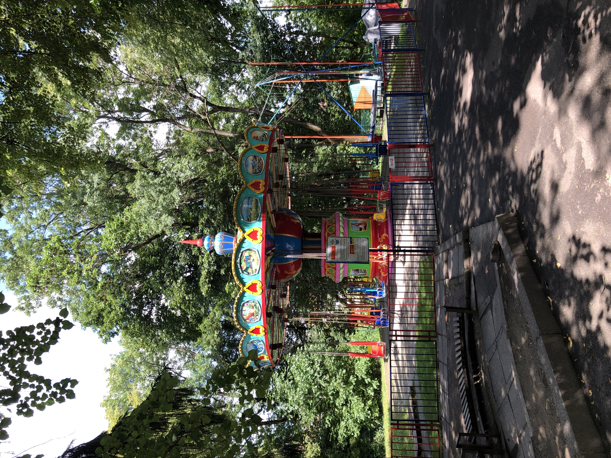 The image shows a merry-go-round amusement ride in a park, likely in Lviv, Ukraine, given the Cyrillic text on a sign. The ride is colorful, with a central structure and suspended seats that resemble swings. It is surrounded by a black and red fence and tall trees, indicating an outdoor setting. The ground is paved with asphalt and has scattered leaves. A park bench is visible in the foreground, partly in shadow. The overall scene depicts a quiet amusement park on a sunny day.