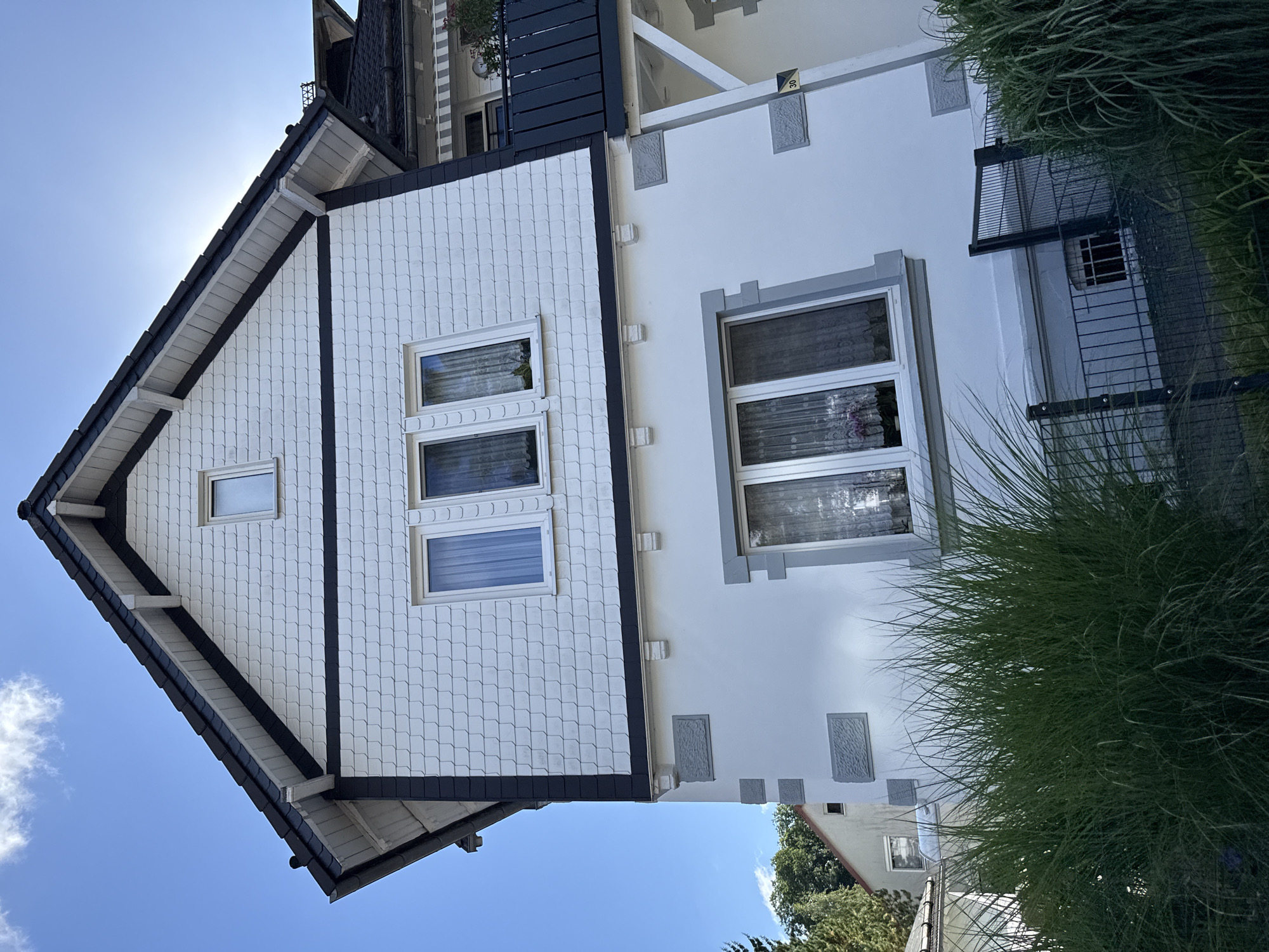 This is a medium-angle, eye-level, outdoor, daytime, vertical, full-color photograph of a white and gray stucco house with a dark gray shingle roof, located in Bad Sachsa, Germany. The house is modern in style and features multiple windows with white frames. The exterior walls are adorned with gray stone-like accents on the lower portion, and the upper portion is covered in white shingles. The roof is steep and dark gray, with white trim along the eaves. In the foreground, tall green ornamental grasses are visible in front of a black metal fence. Behind the main house, another part of a building is visible with a dark gray balcony railing and a white awning. The sky is clear blue with a few wispy clouds, and the sun is shining brightly, casting a glare on the roof. The overall impression is of a well-maintained residential property.