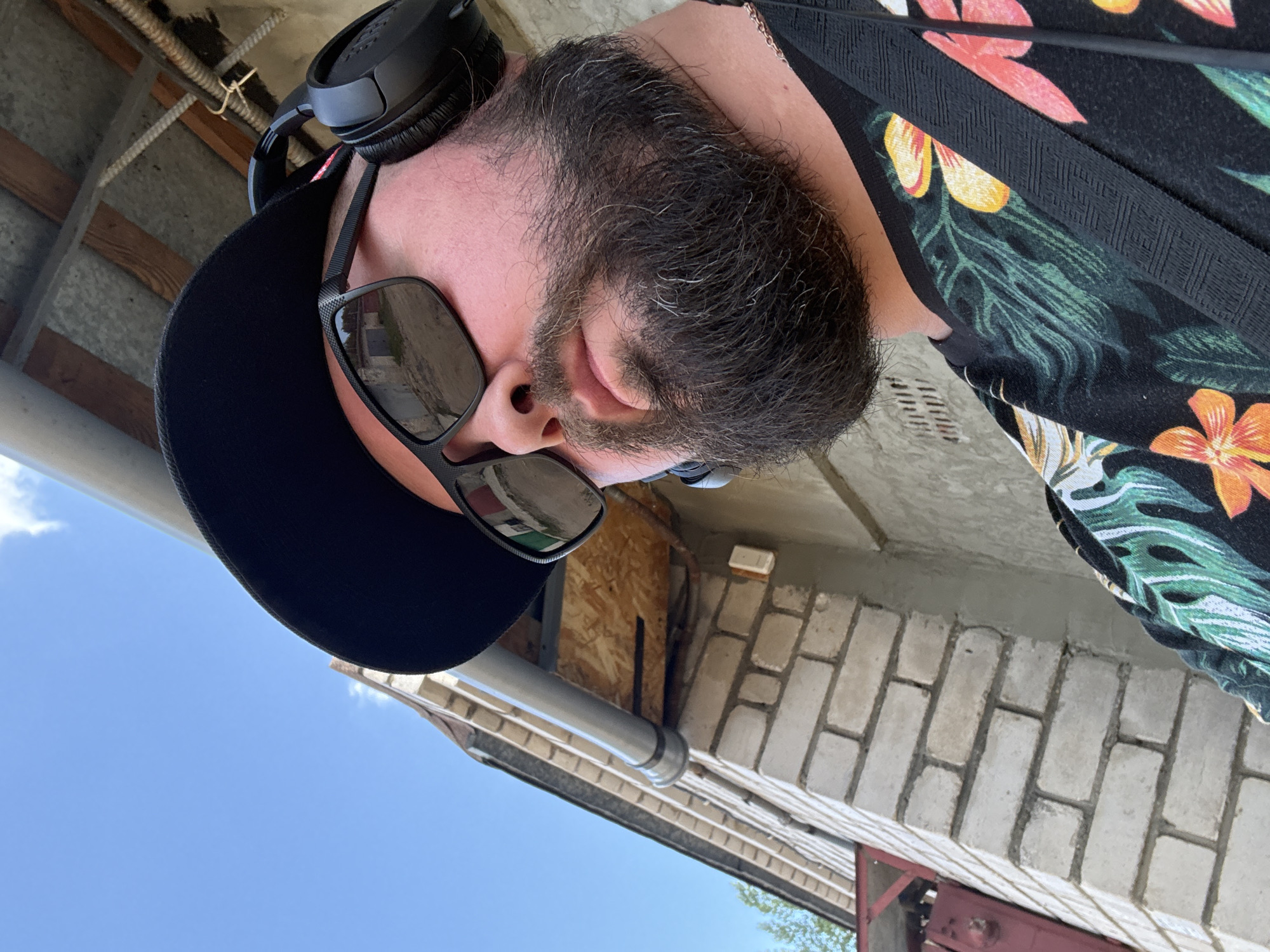 A close-up, low-angle selfie shows a man wearing a black baseball cap, black sunglasses, and black headphones. He has a full beard with some gray hairs. He is wearing a black t-shirt with a colorful floral print. The background consists of a light-colored brick wall, a light gray downspout, and a clear blue sky. Wooden beams and exposed wiring are visible above the downspout. A portion of a red metal structure is on the left side of the image. Based on visual cues like the architecture and building materials, this scene is likely located in Orsha, Belarus.