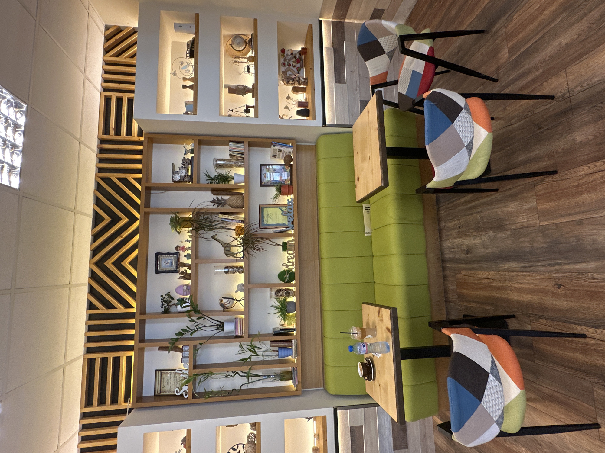 The image shows a cafe interior. A green sofa is situated against a wall with wooden shelving units. The shelves are filled with various decorative items, including plants, picture frames, and globes. Two small wooden tables are placed in front of the sofa. On one of the tables, there is a cup of coffee, a bottle of water, and a straw-equipped drink. In the foreground, there are two chairs with colorful, patchwork upholstery. The floor is made of wooden planks. The walls are decorated with a geometric wooden pattern above the shelves. The overall ambiance suggests a cozy and stylish cafe.