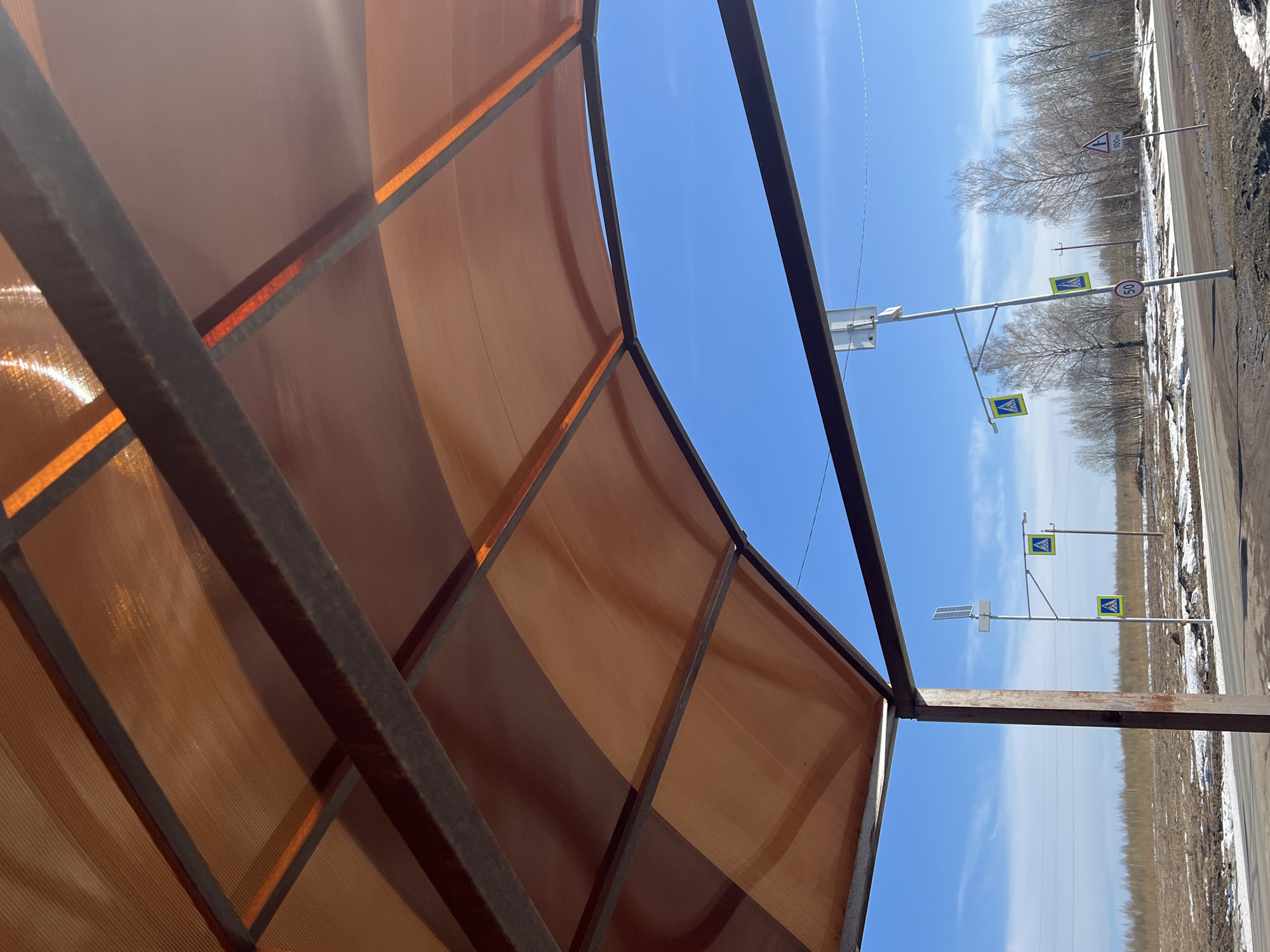 The image shows a view from under a metal and translucent orange polycarbonate structure, possibly a bus stop or shelter, in Gory, Russia. The photograph is taken at an angle, with the ground rotated 90 degrees counterclockwise. Outside the structure, the scene depicts a clear blue sky with some clouds, along with multiple pedestrian crossing signs and a speed limit sign (50). A secondary road intersects the main road with a corresponding road sign (100). There are power lines overhead. Beyond the road signs, there is sparse, dry vegetation and bare trees lining the roadside. Patches of dirt and possibly old snow can be seen near the road.