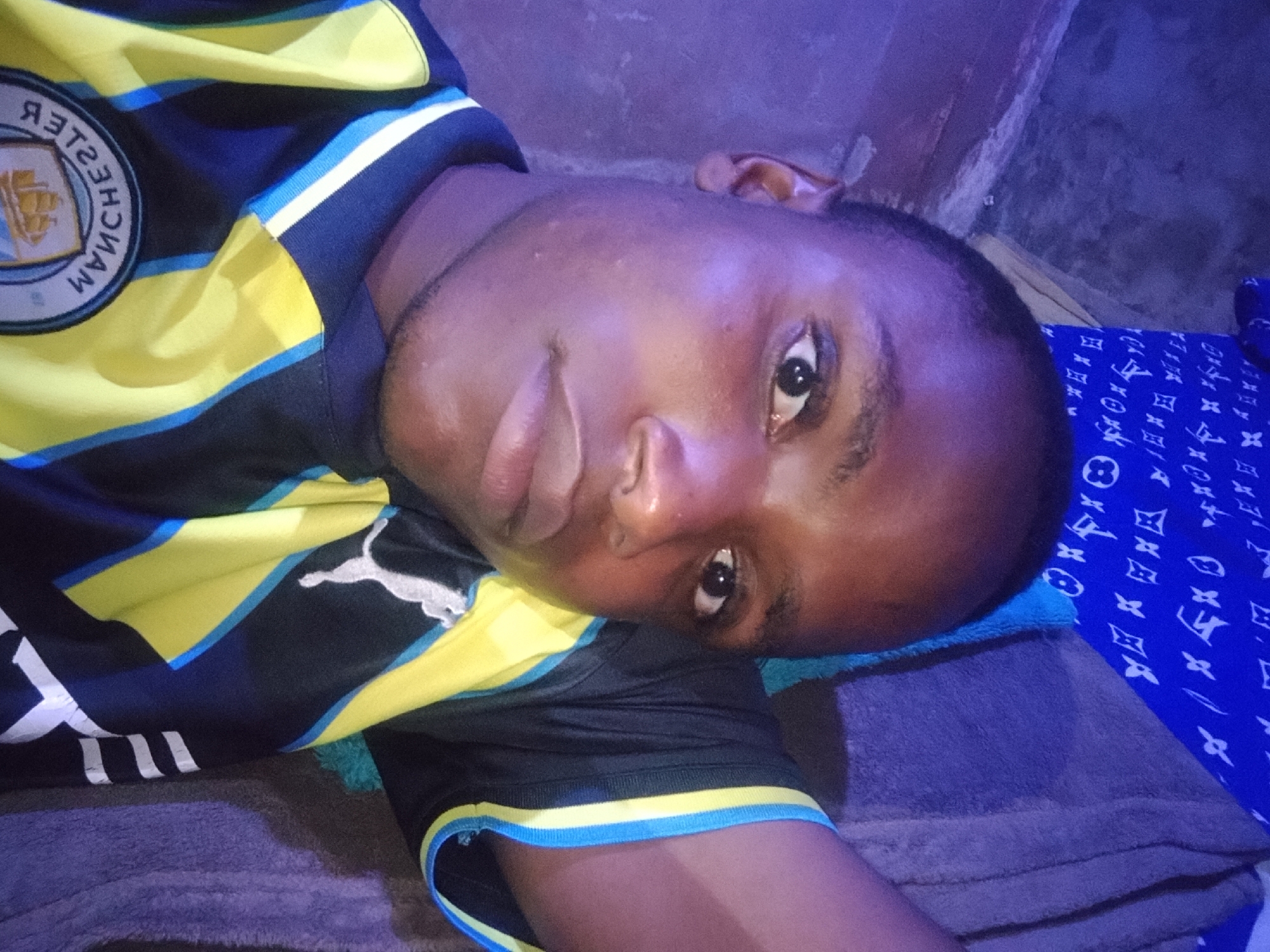 The image shows a close-up, overhead shot of a person lying down, with their face turned towards the camera. The person is wearing a black, yellow, and blue striped jersey with a crest on the left side featuring text and a ship graphic. To the right of the person's head, a blue fabric with a repeating pattern of white logos is visible. The background is a textured, purple surface. The overall lighting is dim and appears to be artificial. The person's skin tone is dark, and they have short, dark hair. The image is slightly blurry, particularly around the edges.