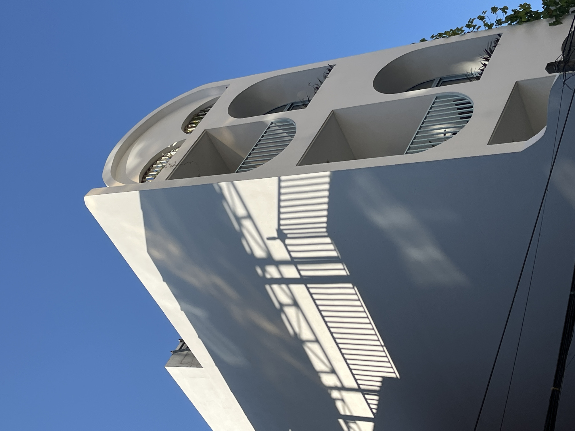 A low-angle shot of a white, modern architectural building against a bright blue sky. The building has a curved top with alternating circular and square cut-outs. Some of these cut-outs have slatted blinds or plants inside. The façade of the building features a prominent shadow pattern resembling blinds or a barred window. Green foliage is visible at the top edge of the building. Based on the architectural style, and the plants, it's likely the image was taken in Đà Nẵng, Vietnam.