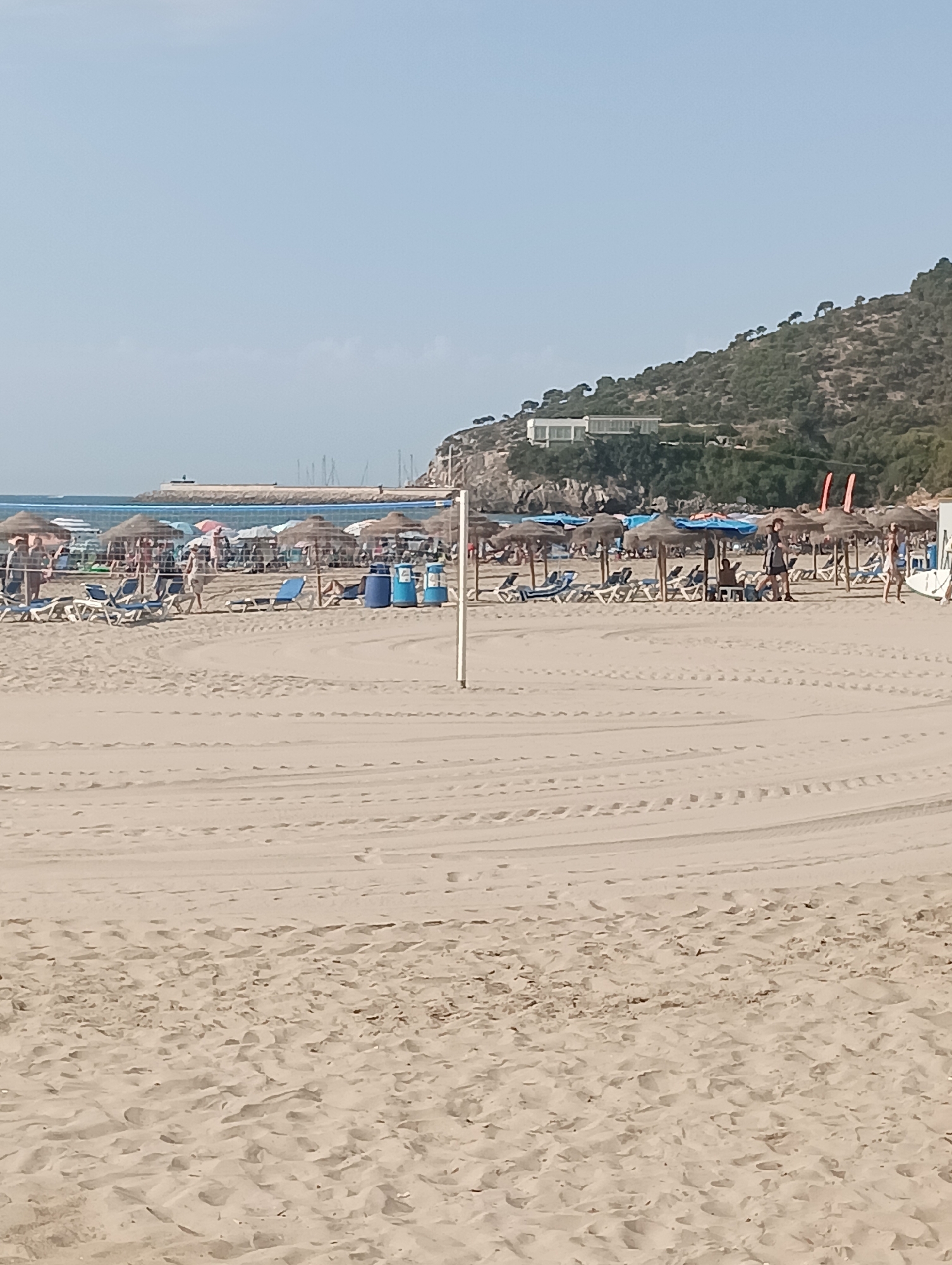 This image depicts a beach scene in La Playa, Spain, with a focus on the sandy expanse leading to the water and a line of beach umbrellas and loungers. In the foreground, tire tracks are visible on the sand, indicating recent vehicle or equipment presence. A white volleyball net pole stands prominently in the center foreground. Behind it, rows of straw-covered umbrellas and blue lounge chairs are arranged, with several people visible scattered among them. Some individuals are standing or walking, while others are seated or reclining. Two blue trash receptacles are placed near the umbrellas. In the background, the ocean is visible with a breakwater or pier extending into the water, and beyond that, a hillside covered with vegetation and a modern building. The sky is clear and pale blue. The overall impression is of a sunny, active beach day.