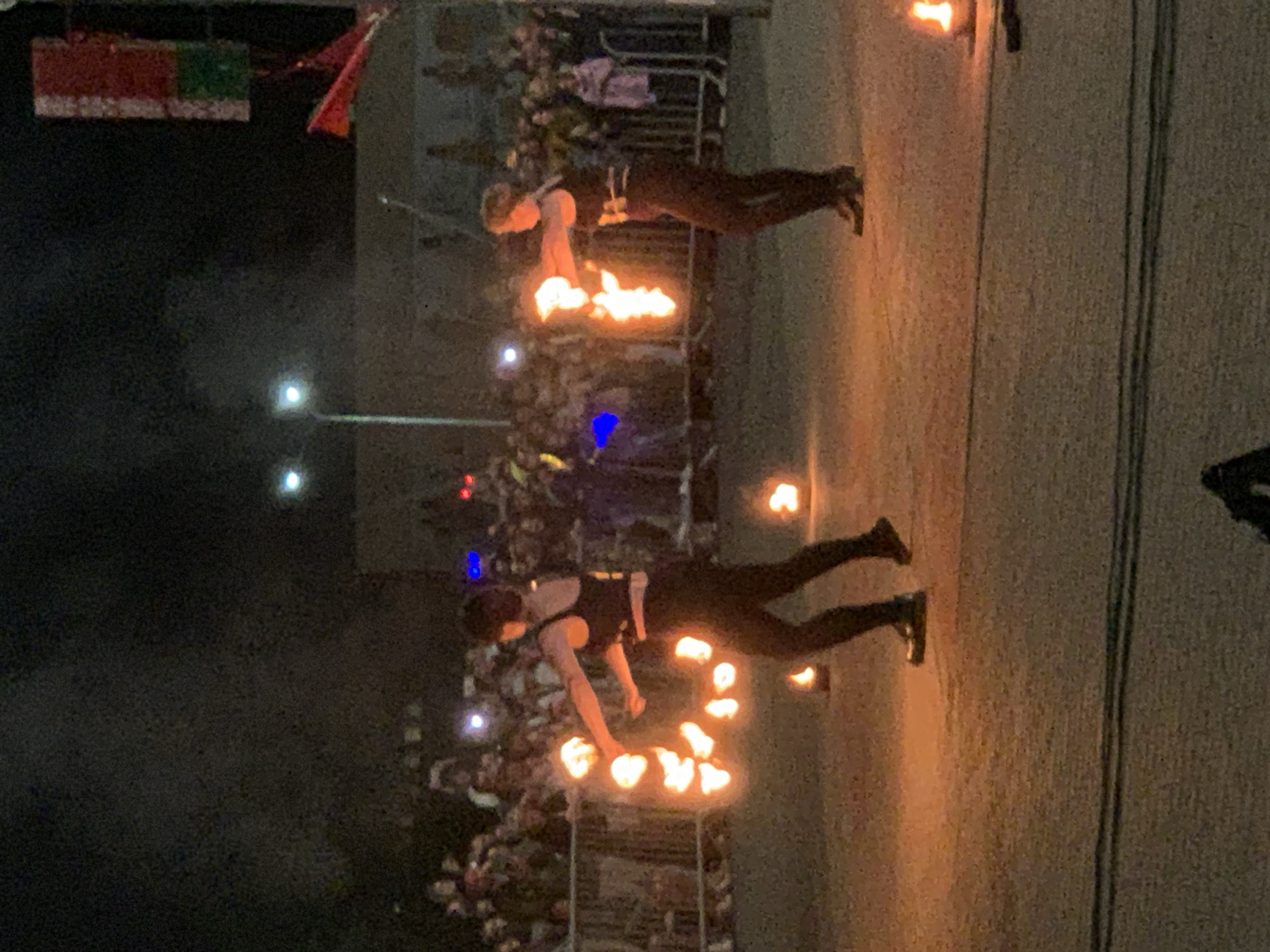 Two individuals are performing with fire at night in front of a crowd. The person on the left is wearing a black sleeveless top and black pants, and is twirling a chain of fire. The person on the right is also wearing a black outfit and is holding a ball of fire in their hands. The ground is illuminated by the fire. In the background, a crowd of people are watching the performance. There are also some lights in the background, including streetlights. A flag with red and green sections is visible in the upper right corner of the image. The scene appears to be outdoors, possibly at a festival or event. The location is identified as Kapyĺ, Belarus.