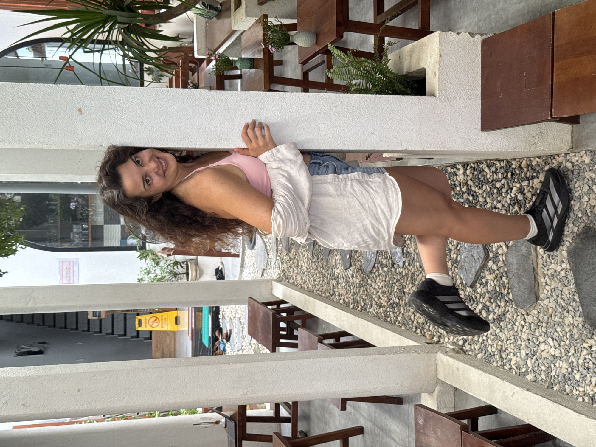 A woman with long, curly brown hair and fair skin is posing for a photo in an outdoor seating area. She is leaning against a white pillar, with her right arm bent and hand resting on the pillar, and her left leg bent at the knee, with her foot lifted slightly off the ground. She is wearing a pink tank top, denim shorts, and black athletic shoes with white socks. Her hair is long and wavy, falling over her shoulders. She is smiling directly at the camera.
The background shows a courtyard with gravel pathways, interspersed with large, flat grey stones. To the left, there are wooden tables and stools, and a set of stairs leading up. A yellow "No Entry" sign is visible near the stairs. To the right, more wooden tables and benches are arranged, some with small potted plants. Lush green plants are also visible in the background, including a large potted plant to the far right. The architecture suggests a modern cafe or restaurant with an open-air design. The overall ambiance is casual and relaxed, typical of establishments found in popular tourist areas like Hoi An, Vietnam.