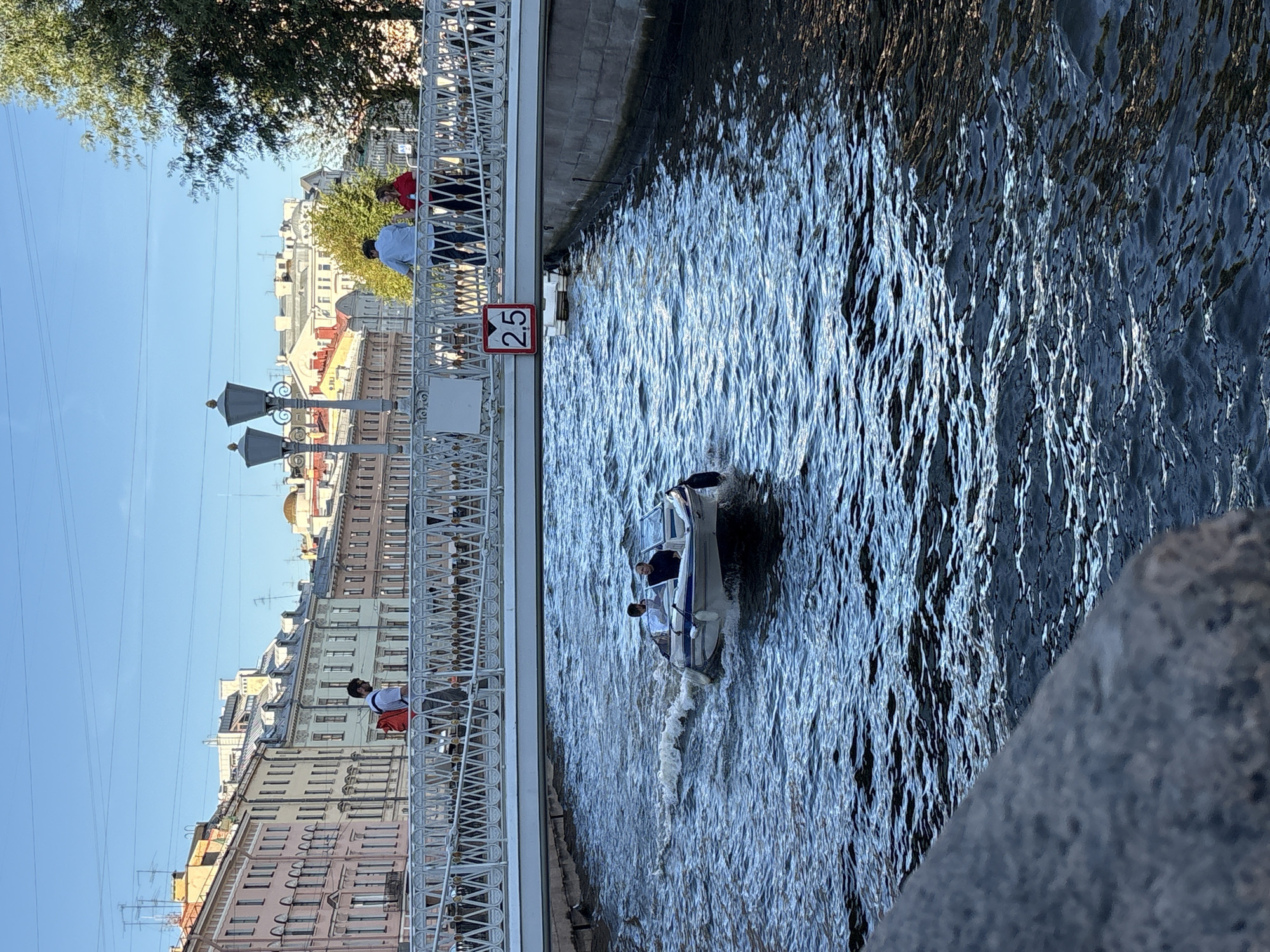 A motorboat carrying two people navigates a canal in Saint Petersburg, Russia. The boat is white with blue trim and is leaving a wake of white foam. The canal is lined with stone embankments. A pedestrian bridge arches over the canal, with people walking across it. Buildings with colorful facades and pitched roofs line the canal banks in the background. A red and white sign with the number "2.5" is visible on the underside of the bridge. The sky is clear and blue. The scene is lit by bright sunlight, casting shadows on the water and buildings.