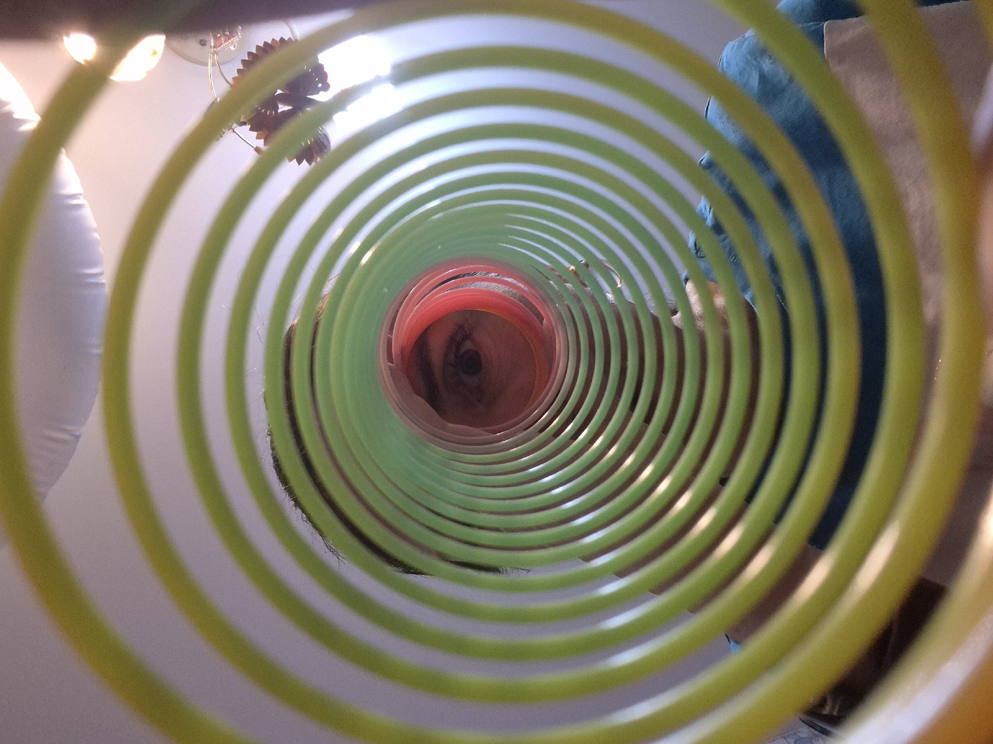 A low-angle shot through a colorful plastic spring toy reveals a person's face peering through the central opening. The toy's coils, in shades of yellow, green, and red, create a concentric ring effect. The person's eye is visible within the red ring. The background shows a ceiling light fixture, a white pillow or cushion, and what appears to be furniture or a wall. The location in the city of Chaikovsky, Russia is unknown. There is no definitive information within the image itself to provide a specific location. The details are limited to what is visible within the frame.