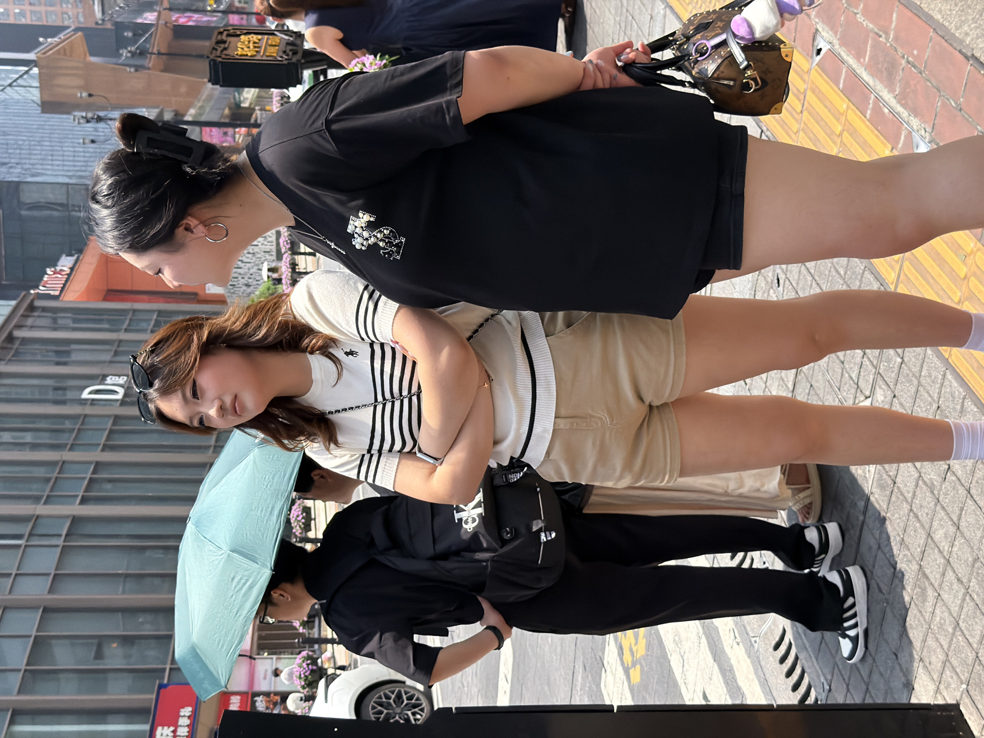 Two young women are standing on a sidewalk in Yuzhong District, China, near a street with a crosswalk. The woman on the left is wearing a striped, short-sleeved shirt and tan shorts, with her arms crossed. She has sunglasses on her head. The woman on the right is wearing a black t-shirt and black shorts, holding a patterned brown handbag. Behind them, a person with an umbrella and a backpack walks away from the camera. In the background, there are buildings and streetlights. The day is sunny.