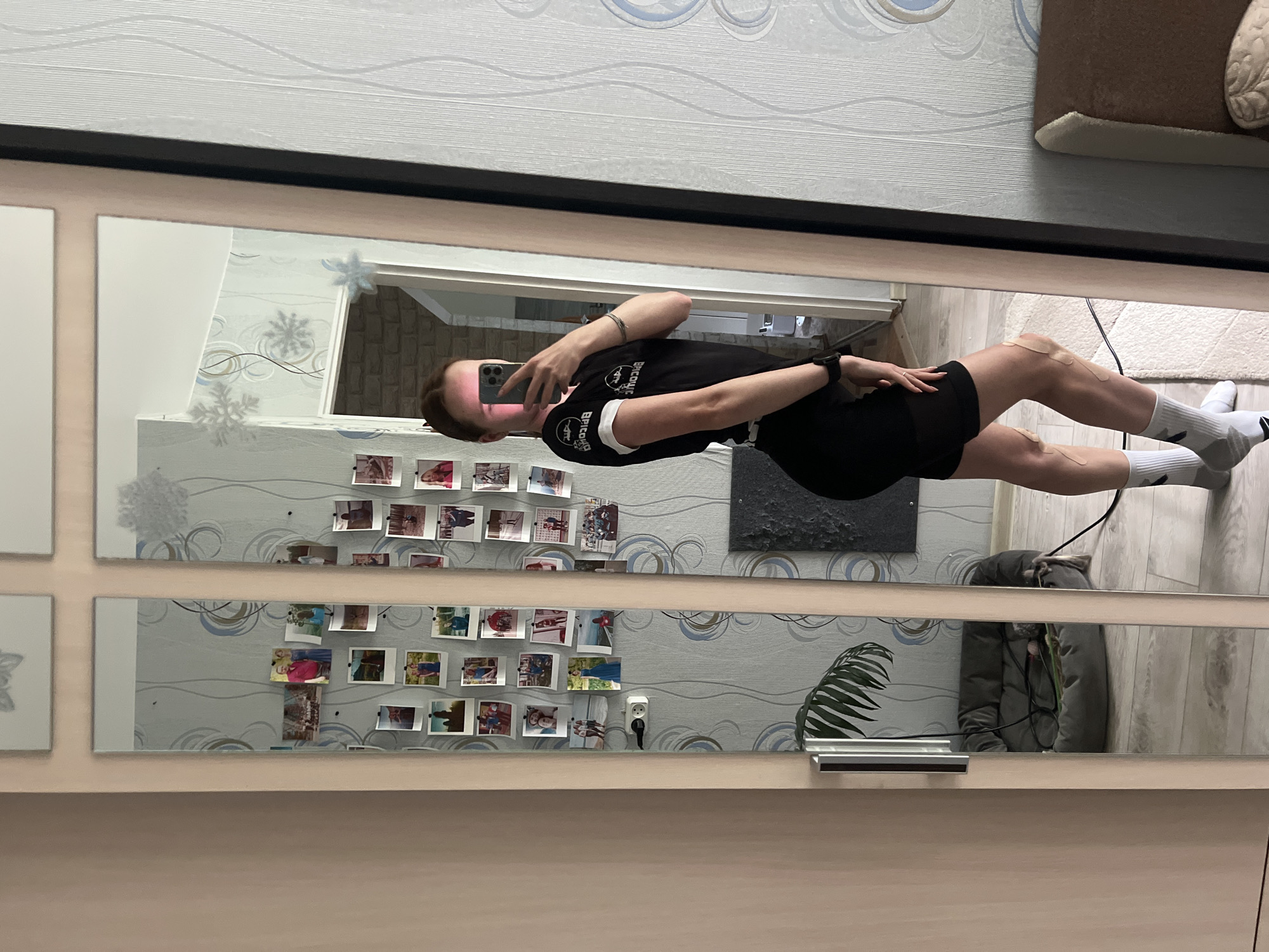 The image depicts a female individual, seen in profile, capturing a mirror selfie within an indoor residential setting in the city of Chaikovsky, Russia. The subject is standing to the right of the central mirror panel, facing left, with her left side toward the viewer. She is wearing a black short-sleeved t-shirt with white trim on the sleeves and collar, displaying reversed white text on the arm that appears to say "BICOML". She is also wearing black athletic shorts, white crew socks with grey accents, and a dark-colored smartwatch on her left wrist. Kinesiology tape strips are applied to both knees. Her face is obscured by a dark-colored smartphone held in her right hand, positioned to capture her reflection.
The mirror she is utilizing appears to be a multi-paneled unit, possibly integrated into a closet or wardrobe, with light-colored wooden or beige frames. The wall behind the mirror is covered with light grey wallpaper featuring a repeating pattern of blue and grey swirls. Numerous small, polaroid-style photographs, depicting various people and scenes, are pinned to this wallpaper. Several white, decorative snowflake cutouts are also adhered to the wallpaper.
Reflected in the mirror, to the left of the subject, a light-colored wall is visible, along with a vertical metallic door handle. Further left in the reflection, a grey cushioned pet bed with a toy inside rests on a light-colored wooden floor. Above the pet bed, a green leafy plant is partially visible. An electrical outlet is positioned on the wallpapered wall. To the right of the subject, another section of the room is reflected, revealing a white patterned door or doorframe and, beyond that, a room with a light brown brick-patterned wall. A portion of brown upholstered furniture with a light-colored, possibly embroidered, armrest is visible in the lower right corner of the reflection, resting on a white textured rug. The flooring throughout the visible areas of the room is light-colored wood or laminate.
