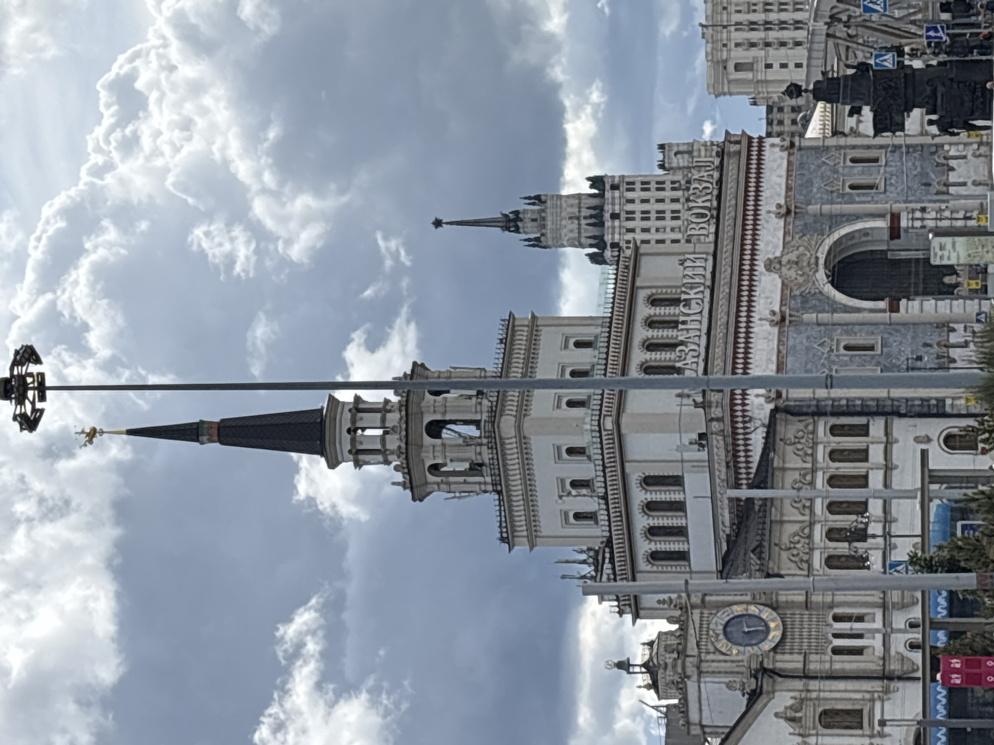 The image depicts the Kazansky Railway Station in Moscow, Russia, as seen from a street-level perspective. The station building showcases intricate architectural details, including turrets, ornate facades, and a prominent clock tower. A portion of the sky, filled with dramatic cumulus clouds, serves as a backdrop. In the foreground, street lighting poles and banners indicate a bustling urban environment. There are no identifiable people visible in the frame. The overall scene captures a blend of historical architecture within a modern city setting.