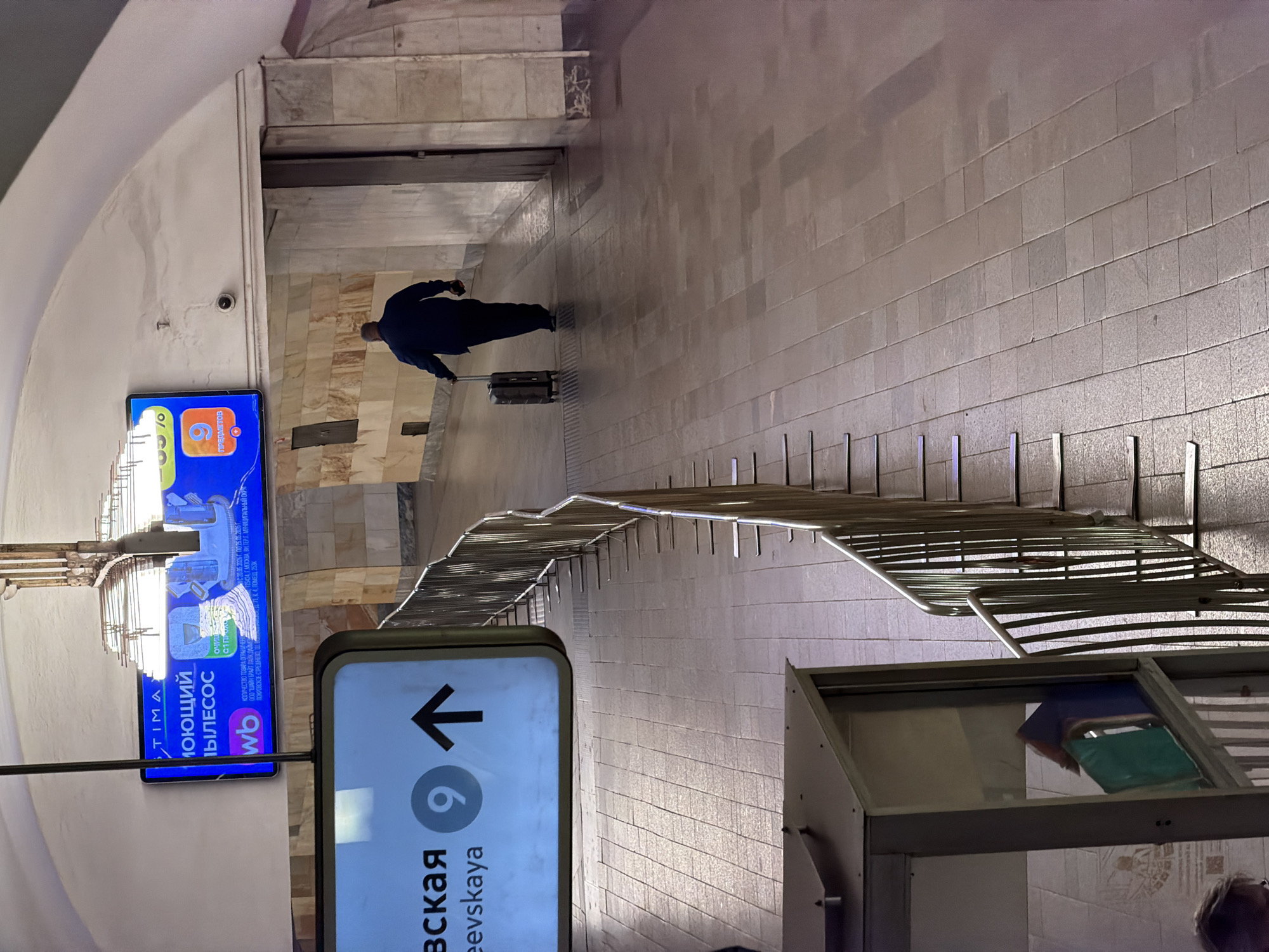 The image shows an interior perspective of a Moscow metro station, most likely the Kiyevskaya station, as indicated by the signage. A person in a dark coat is pulling a suitcase along a corridor. There are railings along the side of the corridor. There is a direction sign on the left showing the number 9 and the name of the station. A blue advertising screen for a product is also visible.