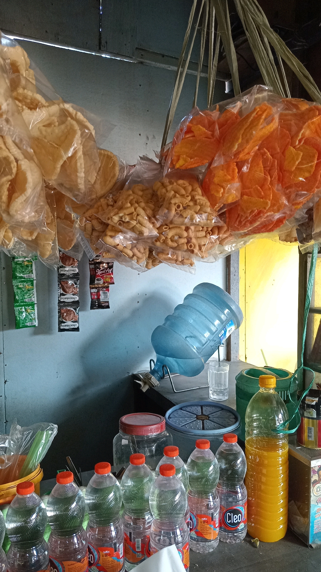 The image depicts a stall likely selling snacks and beverages, possibly located in Ambeng Ambeng Watangrejo, Indonesia. Hanging from the ceiling are several clear plastic bags containing various snacks. One bag holds large, light-colored chips or crackers. Another bag contains smaller, spiral-shaped, tan-colored snacks. A third bag holds bright orange, flat snacks. Below these, on a shelf or wall, are several small packets, some appearing to be for coffee or chocolate.
To the right, a large blue plastic water cooler jug is tilted on a metal stand, with its spigot facing left. A small clear glass is positioned next to it. Further to the right, on the counter, are several plastic bottles. Some contain a clear liquid, while one larger bottle is filled with a bright yellow liquid. There is also a large clear jar with a red lid and a blue plastic container with a circular pattern on its surface. A green plastic tub and various other items are visible in the background. The environment appears to be an outdoor or semi-enclosed space with a rustic aesthetic, indicated by the wooden beams and the simple construction. The lighting suggests it is daytime. No people are visible in the image.