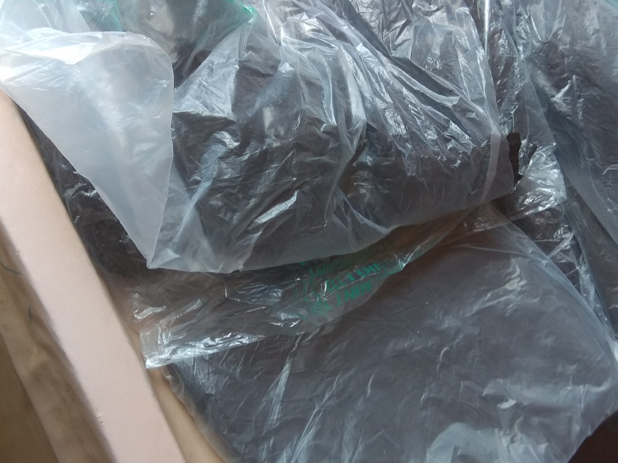The image shows a close-up view of several plastic bags filled with dark material. One bag in the foreground is transparent, revealing the dark contents within. Other bags are partially opaque, obscuring the contents. The bags are wrinkled and appear to be stacked or piled together. The material inside the bags is dark and granular, resembling soil or compost. The overall lighting suggests an indoor environment, possibly a storage area. There are no people or identifiable landmarks visible in the image. The scene appears to be a simple still life of packaged material. There is no information in the image that can indicate its location in Jalingo, Nigeria.