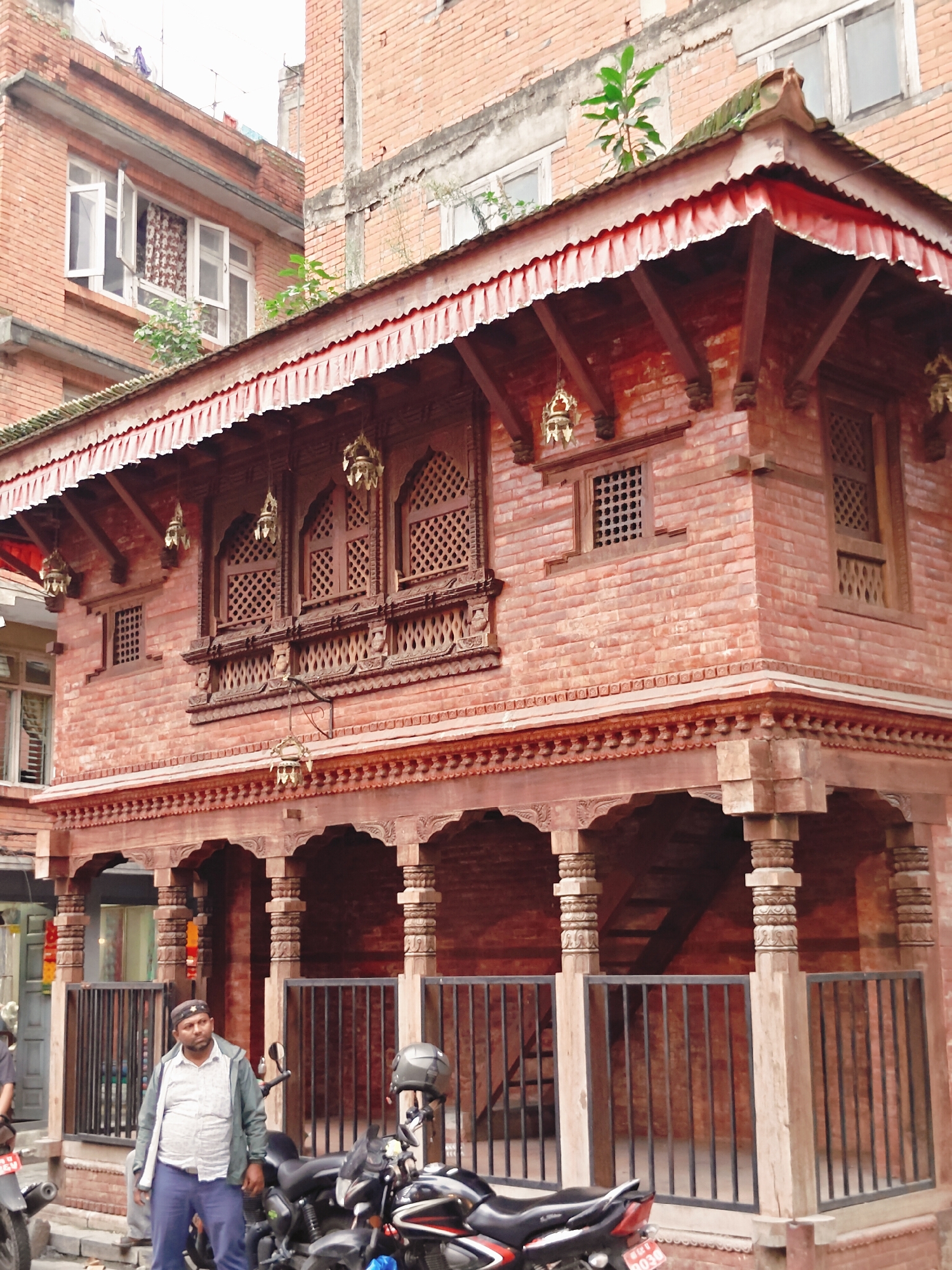 A man stands in front of a traditional red brick building with ornate wooden carvings in Kathmandu Metropolitan City, Nepal. The man, wearing a light-colored shirt, dark pants, and a jacket, is facing the camera. To his right, two motorcycles are parked. The building features multiple levels with intricate windows and decorative wooden elements under a red-tiled roof. The scene suggests a cultural and historical area within the city.