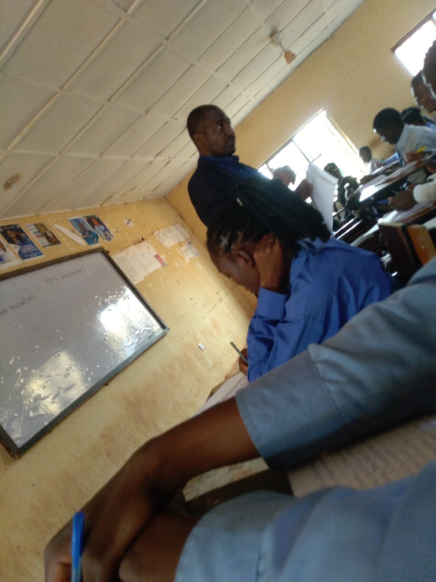 The image depicts a classroom scene, likely in Jos, Nigeria. A teacher, a Black man wearing a blue collared shirt, stands at the front of the room near a whiteboard that has some writing on it. Several students are seated at desks, facing forward. One student, wearing a blue shirt, is leaning on their desk, with their head down. Another student's arm, wearing a blue garment, is prominently in the foreground, holding a blue pen. The classroom appears to be functional, with a tiled ceiling and windows letting in natural light. There are posters or papers affixed to the walls. The overall impression is of a typical educational setting.