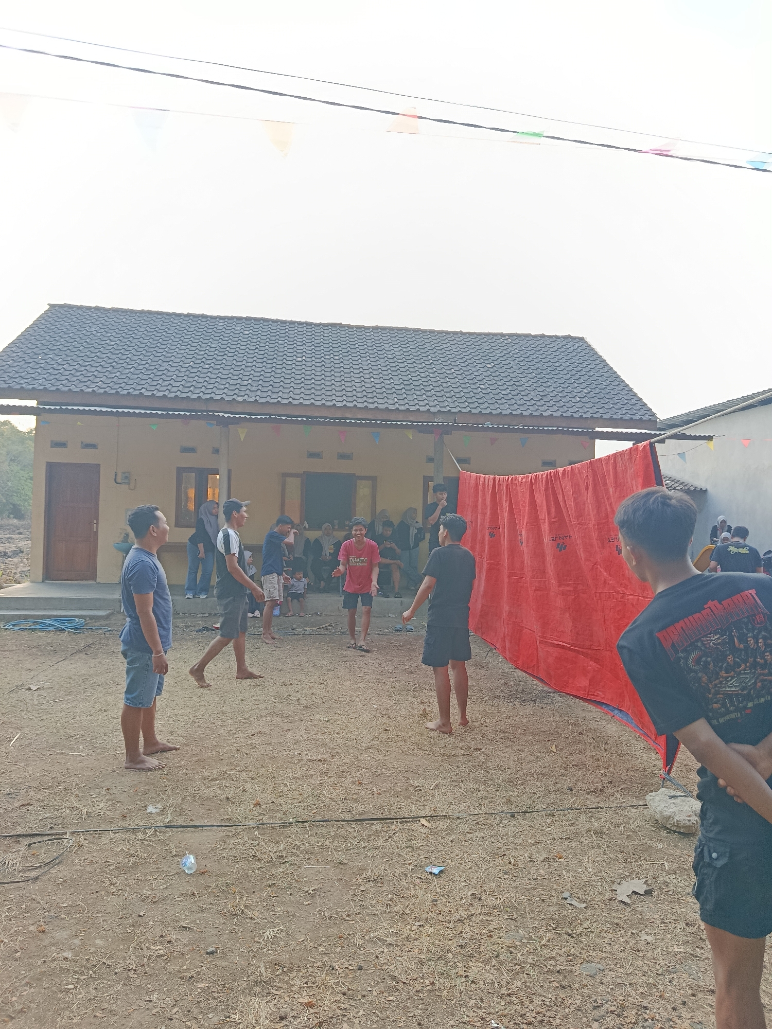 In Sedayu, Indonesia, a group of people are gathered in an outdoor, sandy area in front of a yellow, single-story house with a dark tiled roof. Several individuals, mostly young men, are interacting. Some are standing and looking towards the house, while others are engaged in what appears to be a game, possibly volleyball, with a large red tarp set up as a makeshift net. The tarp has white text on it. In the foreground, a young man in a black t-shirt and black shorts with cargo pockets stands with his back to the camera, facing away from the house and the activity. Another young man in a blue t-shirt and denim shorts is standing to the left, facing the house. The ground is covered in dry grass and dirt, with a black wire running across it. The sky is bright and hazy, suggesting it is daytime. Small, colorful triangular flags are strung across wires above the scene, indicating a possible celebration or event. A few people are visible inside the house through the windows and doorway.