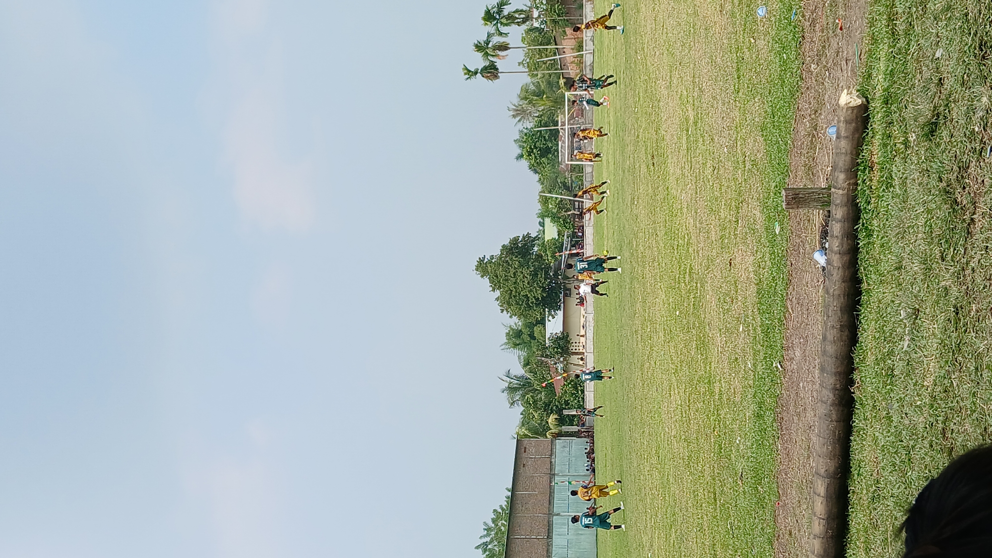A group of young boys are playing soccer on a grassy field under a bright sky. The boys are wearing yellow and green uniforms and are spread across the field, some kicking the ball, others running. In the background, there are buildings, trees, and a goalpost. A large fallen log lies in the foreground, with some plastic bottles scattered on the grass.