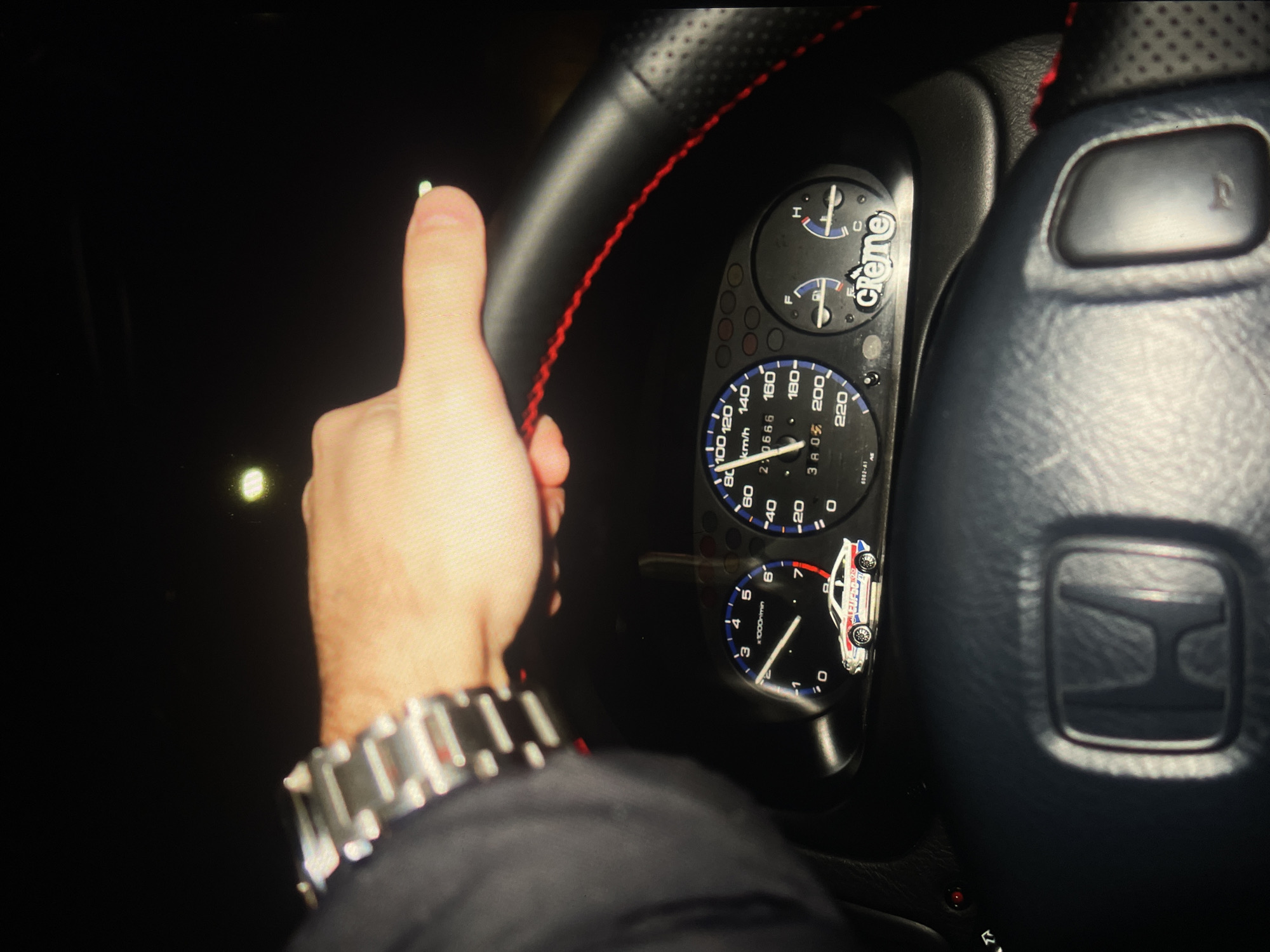 The photograph shows the interior of a vehicle, specifically the driver's side. A hand is visible, giving a "thumbs up" gesture. The driver is wearing a metal wristwatch. The steering wheel is black leather with red stitching. The dashboard is visible, displaying the car's instrument panel, including the speedometer, tachometer, and fuel gauge. A "Creme" sticker is visible near the gauges. A car-shaped sticker is near the tachometer. The car's interior is dark. The scene appears to be illuminated by the dashboard lights. The location is indeterminate.