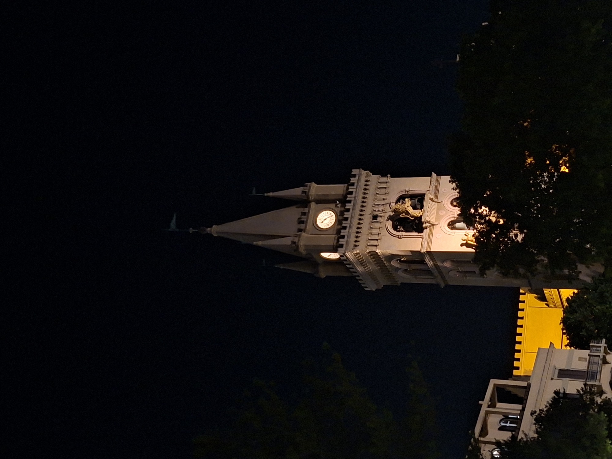 At night, a low-angle shot features a multi-tiered clock tower illuminated with a warm glow. The clock face shows approximately 9:25. The tower is connected to a lower building with yellow-lit walls and a section of a modern-style building with lit windows. Dark trees obscure portions of the scene. The location appears to be Unknown, Unknown.