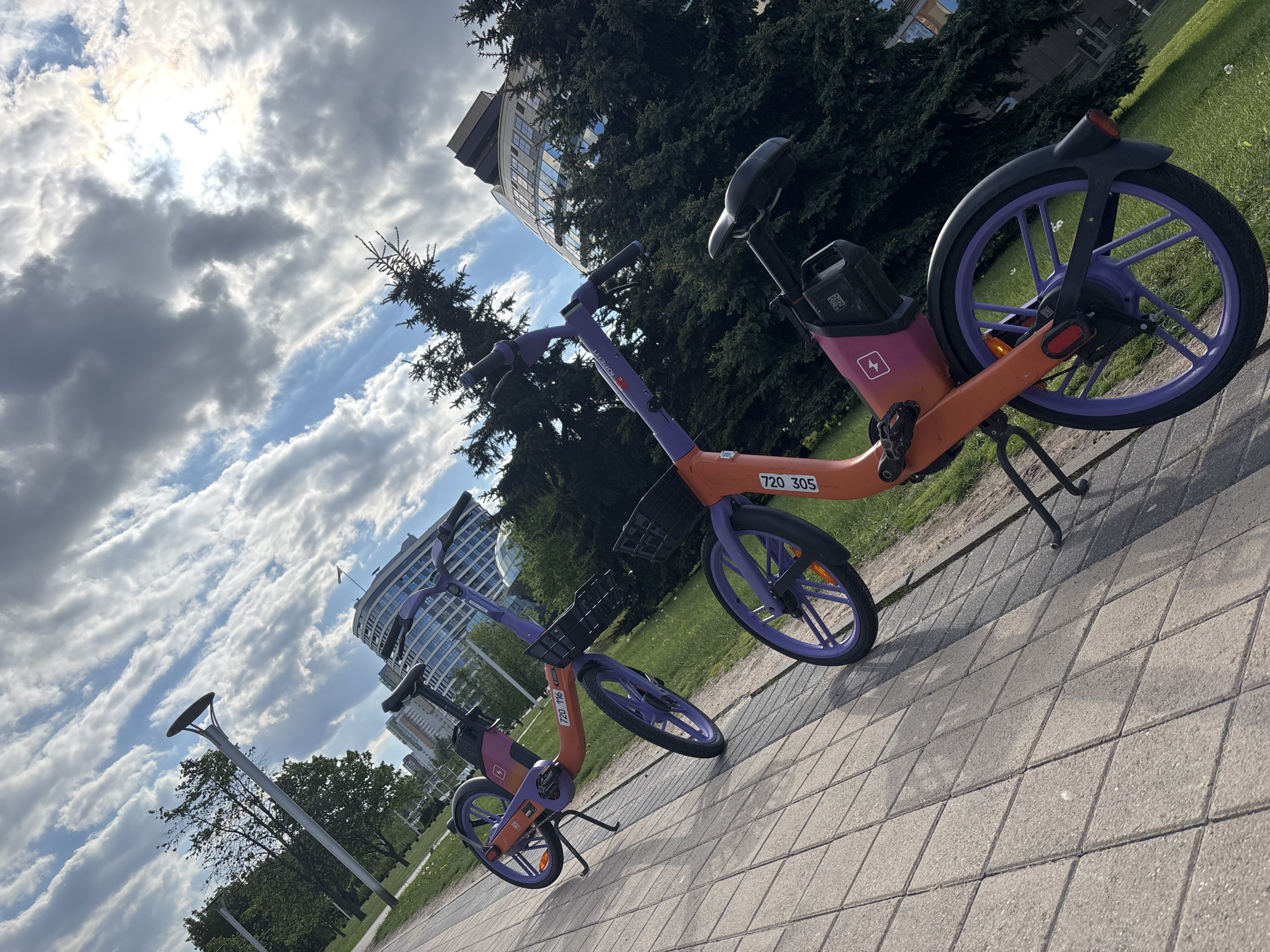 Two orange and purple e-bikes are parked on a paved pathway. The path is made of grey square pavers. Grass and trees are visible in the background, along with a light post and buildings. The sky is filled with clouds and the sun is shining brightly. The e-bikes have baskets in the front and appear to be part of a bike-sharing program. The location is likely in a park or urban area in Minsk, Belarus.