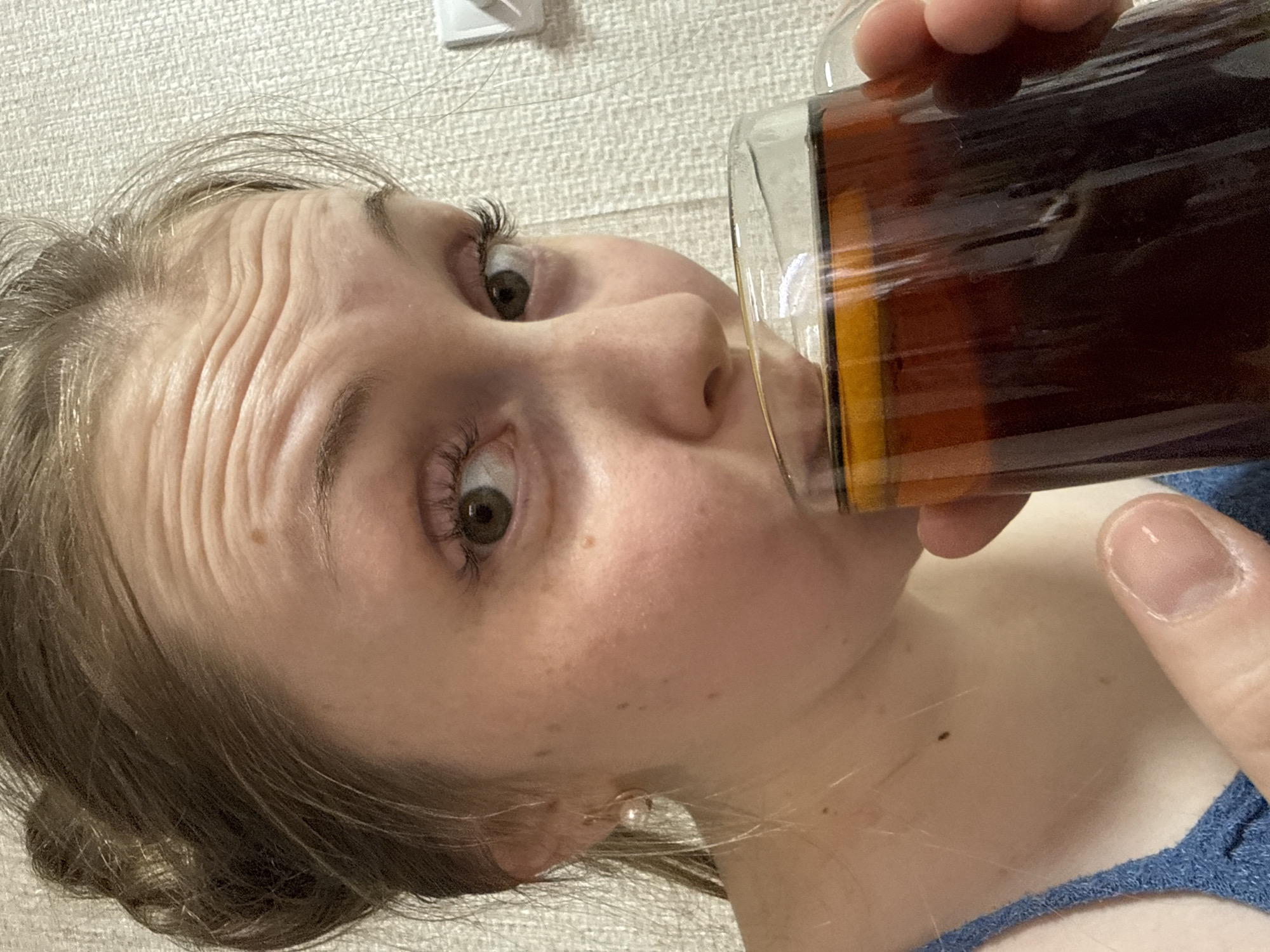 A close-up, rotated image captures a person drinking from a clear glass filled with a dark brown liquid, possibly a beverage like iced tea or soda. The person, who appears to be young with fair skin and light brown hair, is looking directly at the camera with wide, expressive eyes and raised eyebrows, causing prominent wrinkles across their forehead. This suggests a playful, surprised, or exaggerated expression.
The individual's face is central, with a portion of their hair visible on the left and right. They have an earring in their visible earlobe. Their thumb, with a natural, unpolished nail, is seen holding the glass on the right side of the frame, and a glimpse of a blue strap, likely from clothing, is visible near the bottom right. The background features a light-colored, textured wall, suggesting an indoor and casual setting, possibly a home. There are no clear indications of the time of day or specific location. No text is visible in the image.