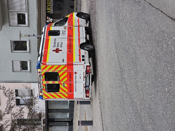 A white German Red Cross (Deutsches Rotes Kreuz) ambulance is parked on a cobblestone surface in Zweibrücken, Germany, specifically in front of Armisan Kebab & Pizza Haus at Hauptstraße 140. The ambulance displays a white body with prominent red and yellow reflective striping, the red cross emblem, and "RETTUNGSDIENST" (Rescue Service) text on its sides and rear. The emergency number "112" is visible on the passenger side, and the license plate "ZW GR 831" is affixed to the rear. Blue emergency lights are mounted on the vehicle's roof.
The scene features a dark gray asphalt foreground leading to the cobblestone area where the ambulance is situated. In the background, a multi-story gray building with several rectangular windows, each fitted with white venetian blinds, houses the "ARMISAN KEBAB & PIZZA HAUS" on its ground floor, indicated by a visible sign. To the left of this building, another structure with a light-colored upper facade and dark ground-floor windows is partially visible, with a bare tree branch extending into the frame from its vicinity. Two dark bollards are positioned on the cobblestone near the ambulance's rear, and a street lamp stands between the ambulance and the gray building. No individuals or ongoing activities are visible.