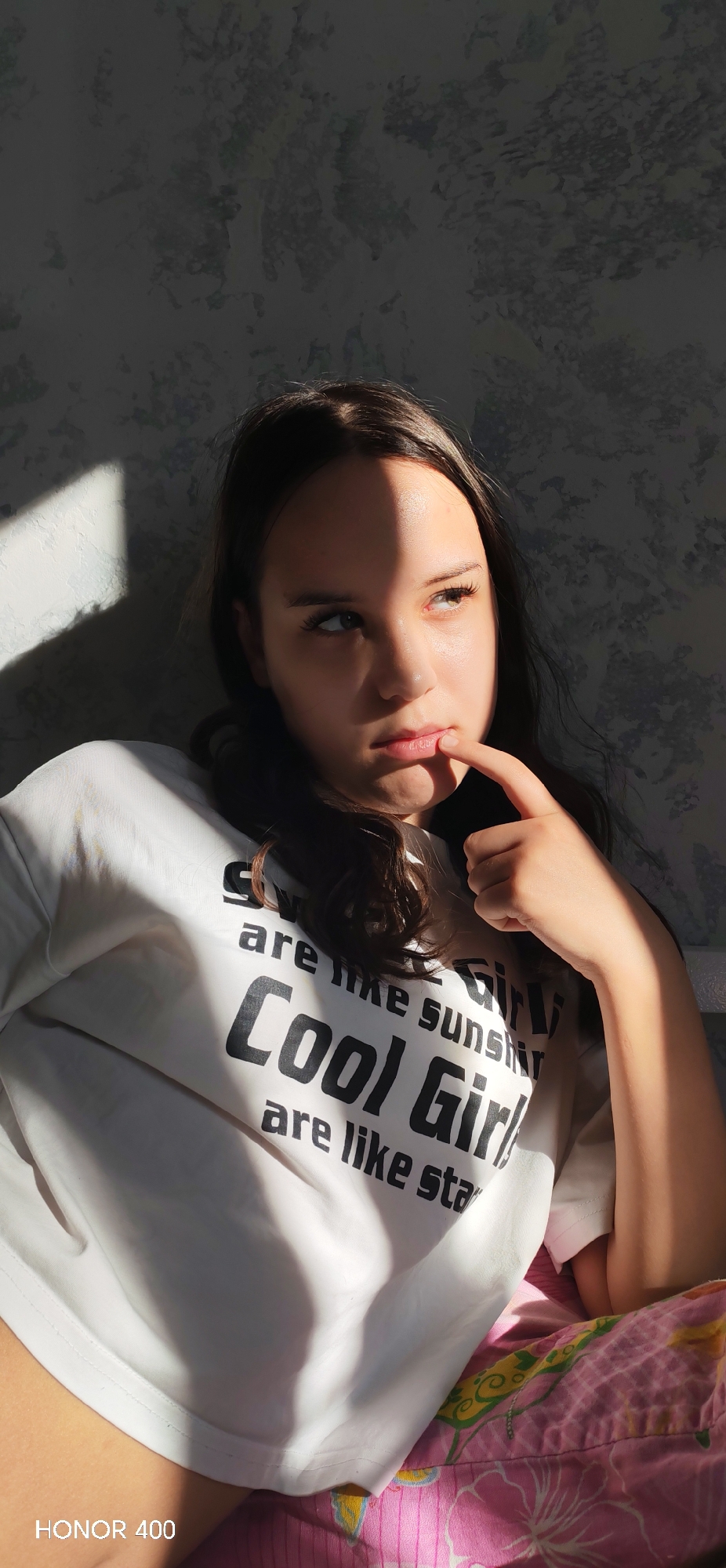 The image depicts a young female individual in an indoor setting. She has dark hair, parted in the middle, and fair skin. She is wearing a white short-sleeved t-shirt with black text that partially reads "are like suns," "Cool Girl," and "are like sta." Her right index finger is positioned on her lower lip, and her eyes are directed towards the upper right of the frame, indicating a contemplative or focused gaze.
A strong, directional light source, likely sunlight, illuminates the right side of her face and body while casting a sharp shadow line across the left side of her face, extending from her forehead down her nose and mouth. A distinct triangular shadow is visible on the wall behind her, to the left. The wall features a mottled, textured pattern in shades of grey. In the lower right portion of the image, a patterned fabric, predominantly pink with yellow and green floral or abstract designs, is visible, possibly belonging to bedding or a cushion. The watermark "HONOR 400" is present in the bottom left corner of the image.
The exact location within Noviy, Russia, cannot be determined from the visual information presented in this image.