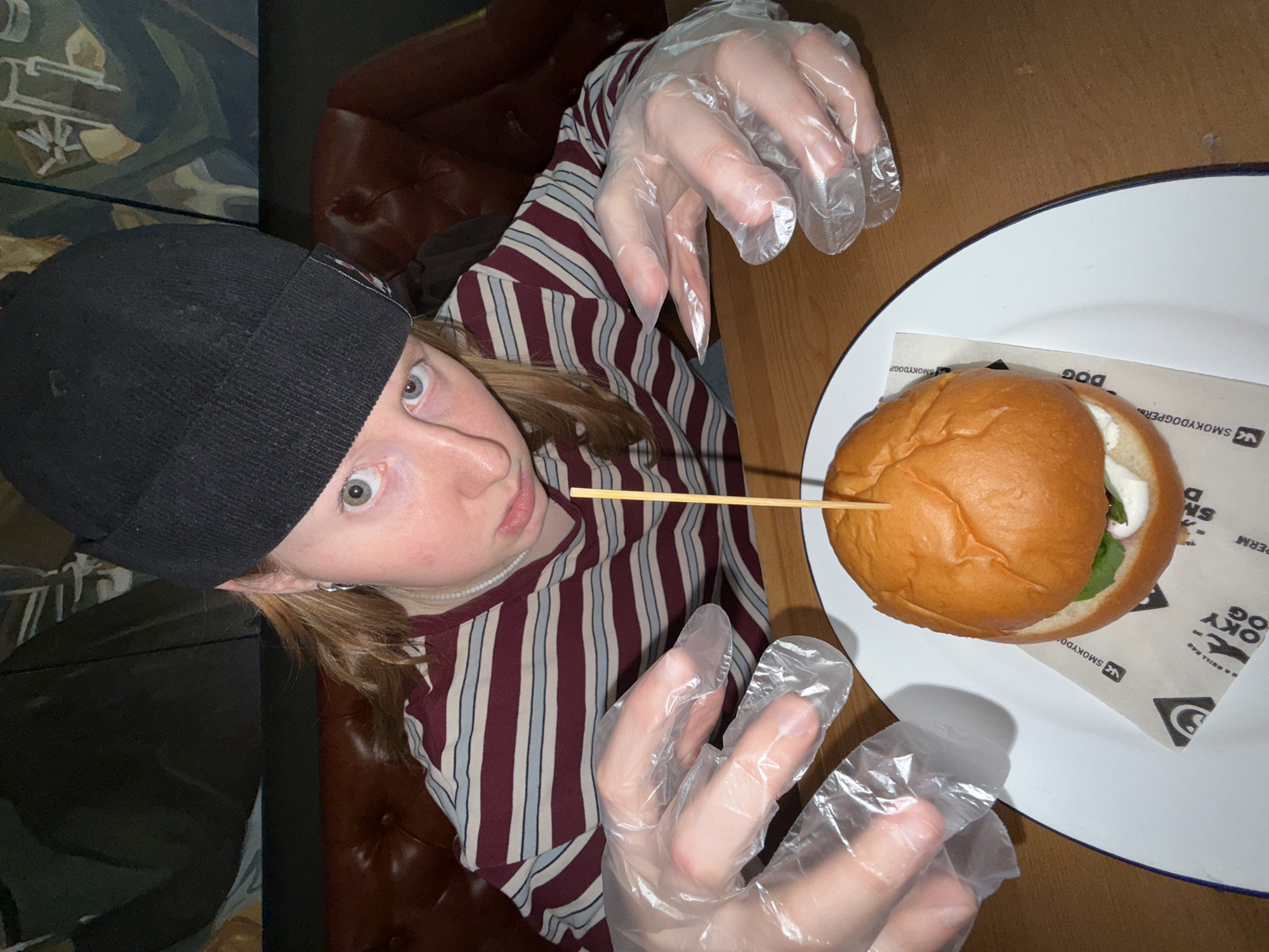 A person with light skin, wearing a black beanie, a pearl necklace, and a maroon and white striped shirt, poses in a selfie with a hamburger on a white plate. The person wears clear plastic gloves on both hands, which are positioned as if reaching for the hamburger. A wooden skewer pierces the hamburger. The plate rests on a wooden table. The logo "SMOKY DOG" is visible on the paper beneath the hamburger. A brown leather chair is partially visible in the background. The location is Perm, Russia.