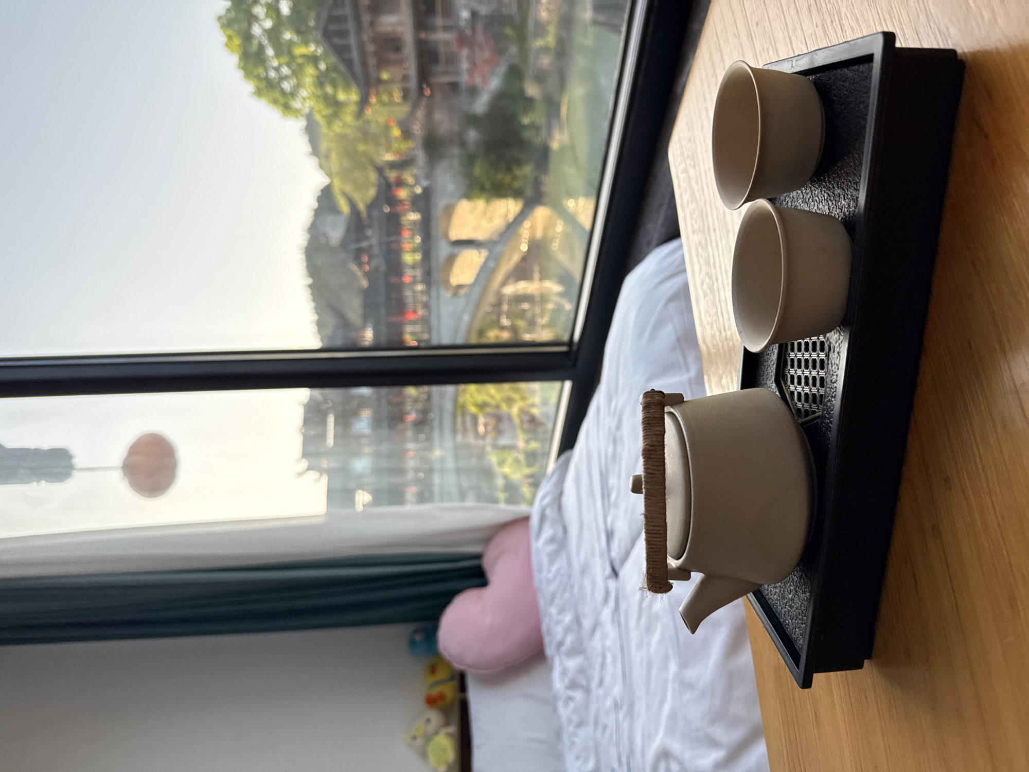The image captures a serene indoor setting with a large window overlooking a scenic traditional town, characteristic of Furong, China. The scene is observed from inside a room, likely a hotel or guesthouse.
In the foreground, on a light wooden surface, a minimalist tea set is presented. It features a light-colored, possibly ceramic, teapot with a textured, rope-wrapped handle, accompanied by two matching teacups, all resting on a black rectangular tray with a patterned, possibly draining, surface. Adjacent to the tea set, a neatly made bed with white bedding and a soft pink heart-shaped pillow is partially visible. Further down, near the bottom of the window, a dark green or blue curtain is drawn, and several small, colorful plush toys can be seen.
Through the expansive window, a bright outdoor vista unfolds. Traditional Chinese buildings with intricate roofs and some red accents are nestled among verdant trees. A distinctive bridge structure is also visible, hinting at a river or waterway flowing through the town. The lighting suggests daytime, with bright sunlight creating a slight haze and a prominent reddish, circular reflection, possibly of the sun or an interior light source, in the upper left portion of the windowpane.
The overall impression is one of calm and relaxation, inviting a moment of rest while enjoying the picturesque view. There are no people or visible text in the image.