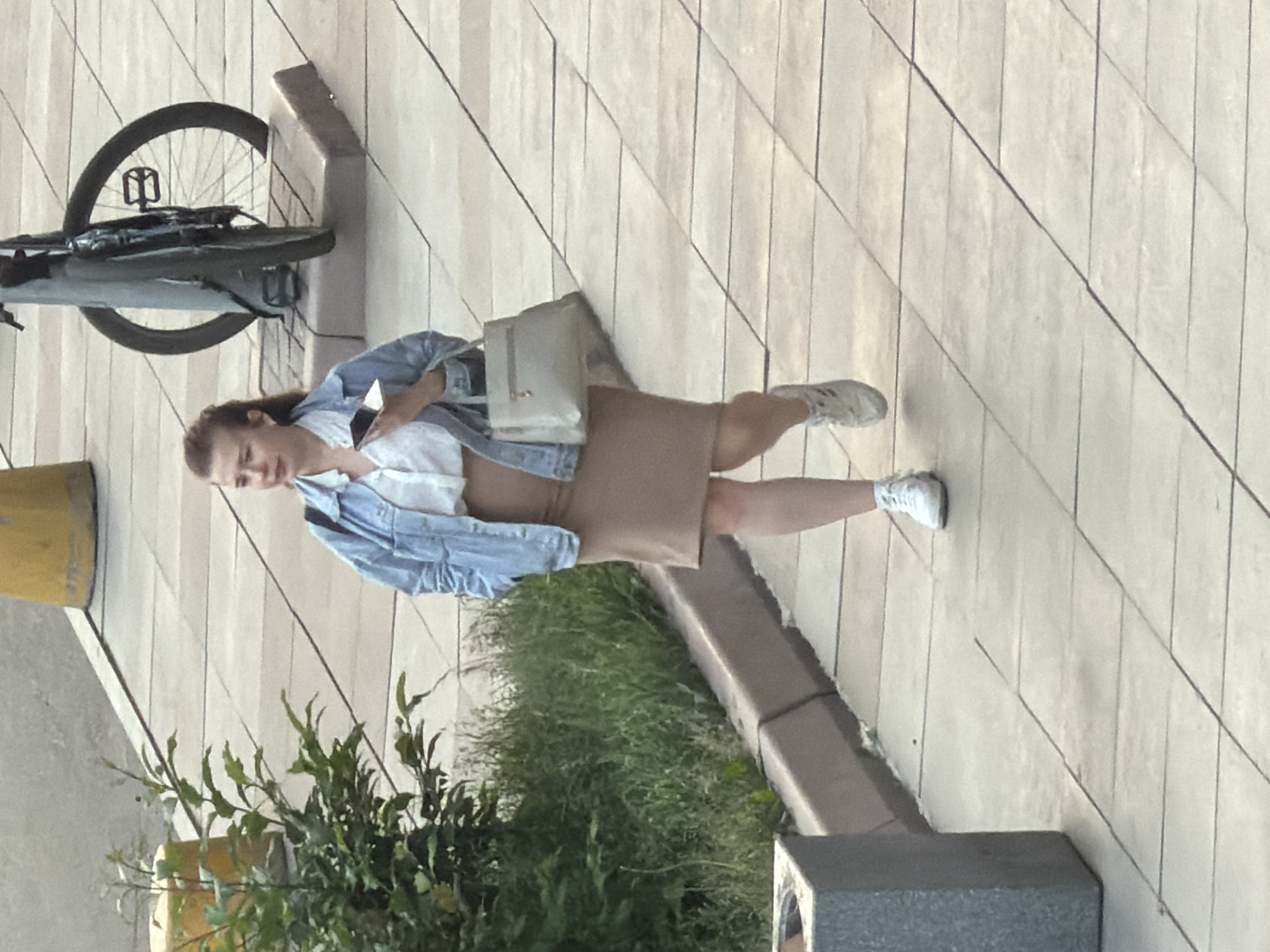 In this media file, a woman is walking on a paved, light-colored outdoor surface, possibly a sidewalk or plaza, composed of rectangular tiles laid in a diagonal pattern. She is wearing a light blue denim jacket over a white top and a light brown skirt. White sneakers are on her feet, and she carries a light-colored shoulder bag. In her right hand, she holds a smartphone. Her hair is dark brown and pulled back.
To her left, a bicycle with a dark frame and a black wheel is parked upright. The background shows a low-lying green hedge and some larger green foliage, suggesting an urban green space. The scene is illuminated by natural light, indicating it is daytime. The overall mood appears casual and everyday. The location is identified as Krasnoyarsk, Russia.