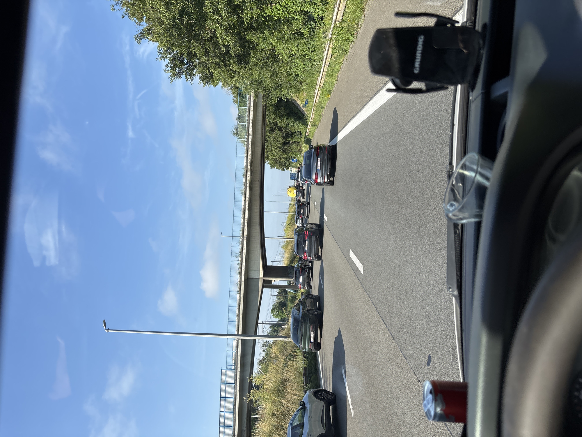 The image is taken from the perspective of a vehicle driver on a highway, likely in or near Vilvoorde, Belgium, given the presence of a street sign with what appears to be Belgian license plates and European highway infrastructure. The scene depicts moderate traffic with several cars, mostly black SUVs and sedans, driving on multiple lanes. An overpass crosses the highway in the mid-ground, with grassy verges and trees lining the sides. The sky is clear blue with some wispy clouds. In the foreground, on the dashboard of the vehicle, is a red can and a phone holder with the brand "GRUNDIG" visible. The overall impression is that of a normal daytime commute on a highway.