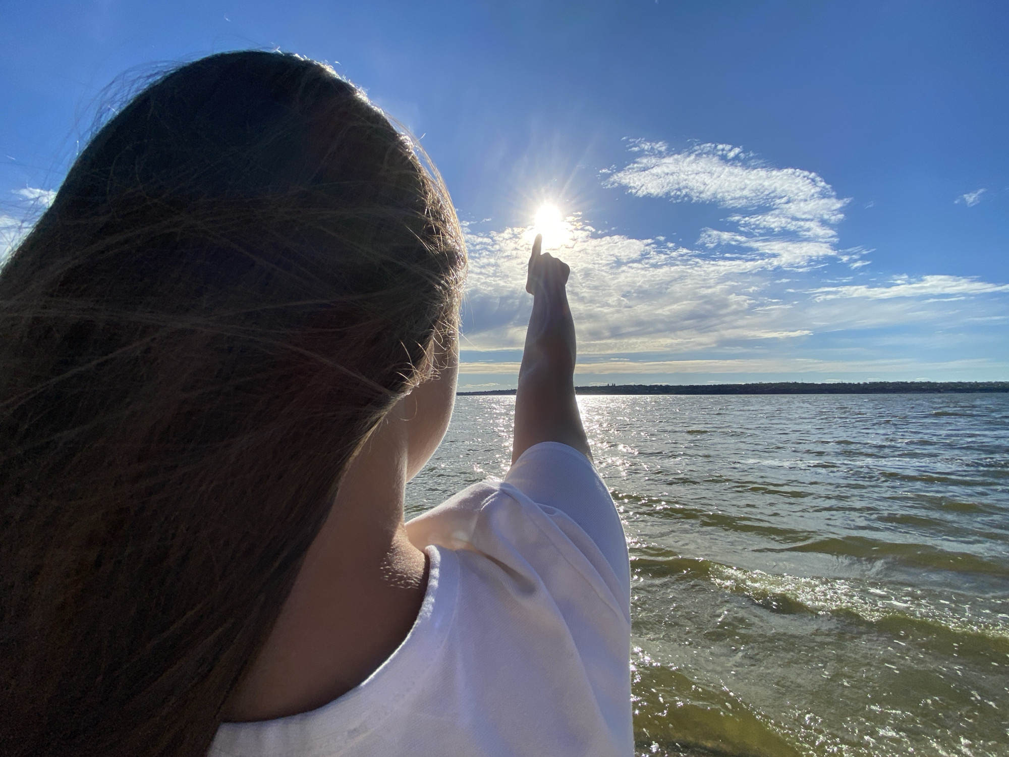 A person is shown from the back, with their right arm extended upwards and their index finger pointing towards the sun. The sun is bright and positioned in the sky, partially obscured by clouds, creating a lens flare effect. The person has long, dark hair that is blowing in the wind. They are wearing a white t-shirt. The background shows a large body of water, likely a lake or ocean, with gentle waves reflecting the sunlight. The sky is blue with scattered white clouds. The horizon line is visible in the distance, with a faint silhouette of what appears to be a cityscape or landmass. The overall setting suggests an outdoor location by the water on a sunny day.
