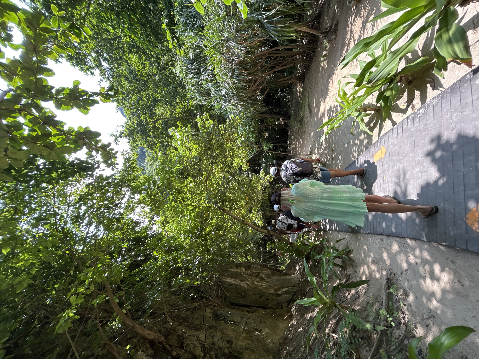 A group of people walk along a raised boardwalk through a lush, tropical environment. The boardwalk is made of dark gray wooden planks and is bordered by sandy ground and vegetation. To the left, a sheer rock face is visible, covered in vines and moss. To the right, a dense canopy of green trees and plants creates a shaded pathway. Sunlight filters through the leaves, casting dappled shadows on the ground. The people are walking away from the camera, their backs to the viewer. The person in the foreground, wearing a light green cover-up, is the most visible. The environment suggests a natural park or a trail leading to a scenic location, likely within the vicinity of Ao Nang, Thailand, given the tropical foliage and rocky terrain.