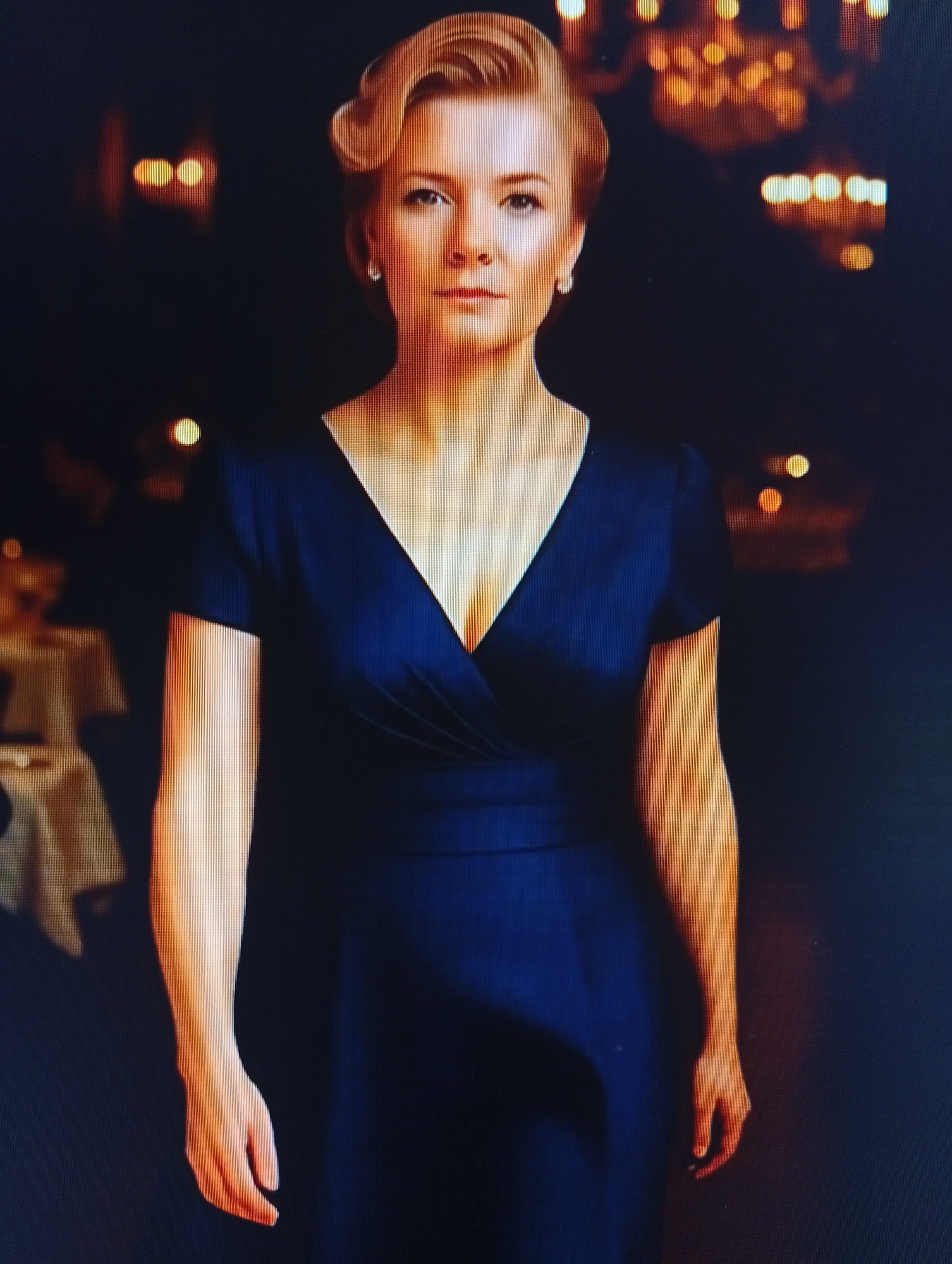 A full-body, medium shot photograph captures a woman from the chest up, facing the viewer directly. She has blonde hair styled in an updo with curled bangs framing her face. Her eyes are open and looking forward. She wears a dark blue, V-neck dress with short sleeves. A pearl earring is visible on her right earlobe. Her arms are positioned by her sides. The background is dark and out of focus, with blurred, warm-toned lights suggesting a formal indoor setting, possibly a restaurant or ballroom, with tables covered in white cloths visible on the left. The lighting accentuates the woman's face and arms. The image appears to be a photograph taken of a screen.
