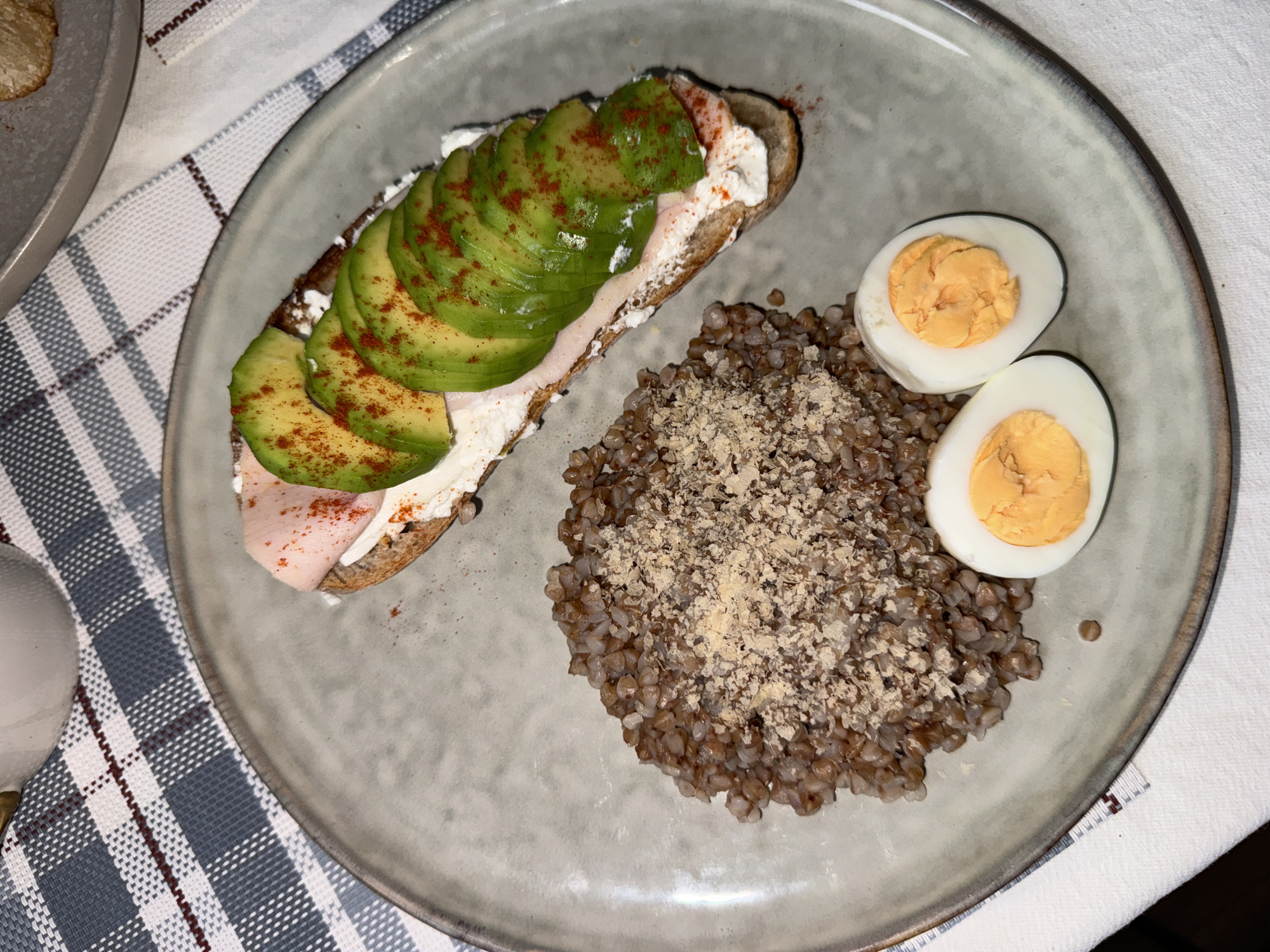 The image presents a top-down, slightly angled view of a prepared meal served on a single, round, light-grey ceramic plate with a darker, irregular rim. The plate occupies the majority of the frame.
The meal consists of three distinct components:
1. **Buckwheat:** A mound of cooked, brown buckwheat groats is situated in the lower-left to mid-left portion of the plate. It is topped with a generous sprinkling of a light brown, granular substance, consistent with finely ground nuts or a seasoning mix.
2. **Hard-boiled Eggs:** Two halves of a hard-boiled egg are positioned in the lower-right section of the plate. Both halves display firm, yellow-orange yolks surrounded by solid white albumen.
3. **Open-faced Sandwich/Bruschetta:** An elongated piece of dark bread, possibly a baguette slice, runs diagonally across the upper-right section of the plate. It is spread thickly with a white, creamy substance, likely cream cheese or a similar dairy spread. Layered on top are two folded slices of light pink deli meat (e.g., ham or turkey), which are then covered with approximately seven uniformly sliced, crescent-shaped pieces of green avocado. The avocado slices and parts of the sandwich are dusted with a fine red powder, consistent with paprika or a mild chili spice.
The plate rests on a tablecloth featuring a checkered pattern of alternating light blue-grey and white squares, accentuated by thin dark red lines forming part of the grid. A plain white textile is visible underneath and to the right of the checkered tablecloth. In the upper-left corner, the rim of a white ceramic mug or cup is partially visible. The edge of another grey plate or dish can be seen in the upper-right background.
No individuals are present in the image. No specific actions or interactions are depicted beyond the presentation of the meal. The environment appears to be an indoor dining setting. There are no visual cues such as landmarks, distinctive architecture, or text that would enable the identification of the specific city as Krasnoyarsk, Russia. The meal and setting are generic.