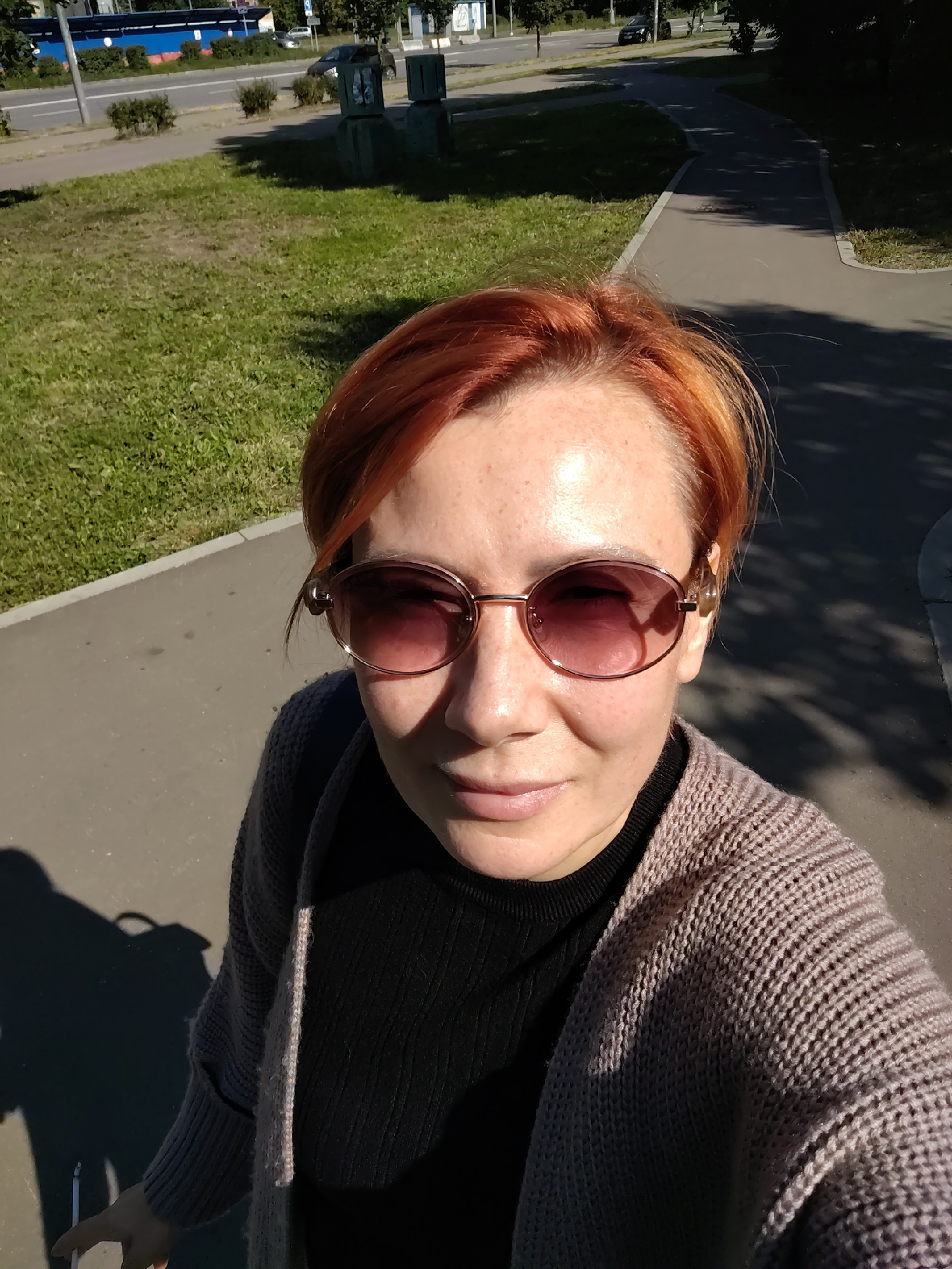 A woman with short, reddish-orange hair and freckles is taking a selfie outdoors in Zelenograd, Russia. She is wearing large, oval, rose-tinted sunglasses and a dark turtleneck sweater under a light brown knitted cardigan. The background shows a paved walkway bordered by a grassy area, a street with a car, and some trees. The sun is casting a strong shadow of the woman and what appears to be a bag on the walkway. The overall impression is a casual, sunny day in a city park or residential area.