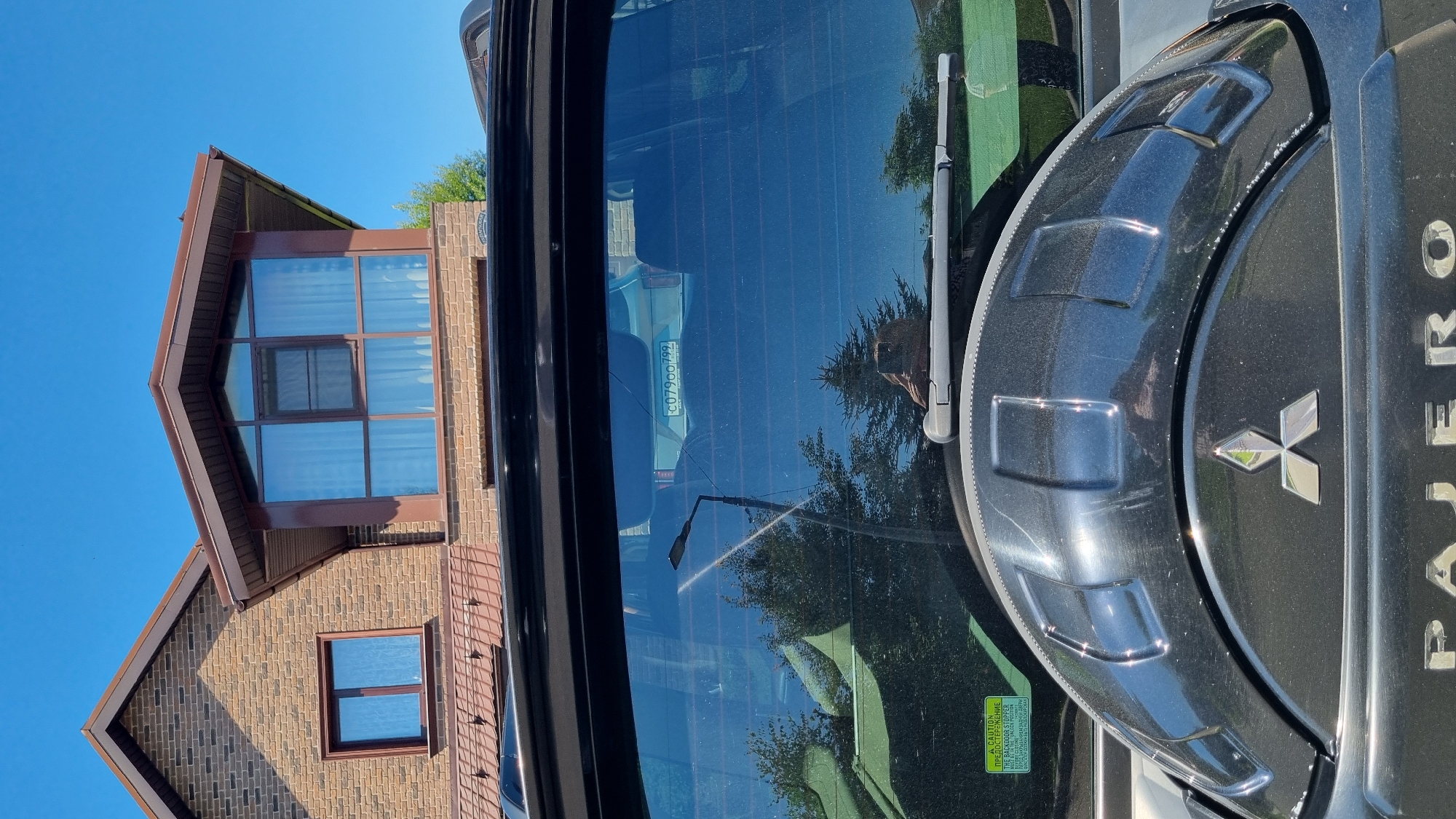 In the city of Verzilovo, Russia, a black Mitsubishi Pajero is parked in front of a two-story brick house with a brown roof. The house has multiple windows, including a large bay window on the second floor. The car's rear window is visible, reflecting the sky and trees. A license plate with the characters "C07900 799" is affixed to the car. The spare tire cover, emblazoned with the Mitsubishi logo, is prominent on the back of the vehicle. A caution sticker is visible on the lower left side of the rear window. The sky is clear and blue.